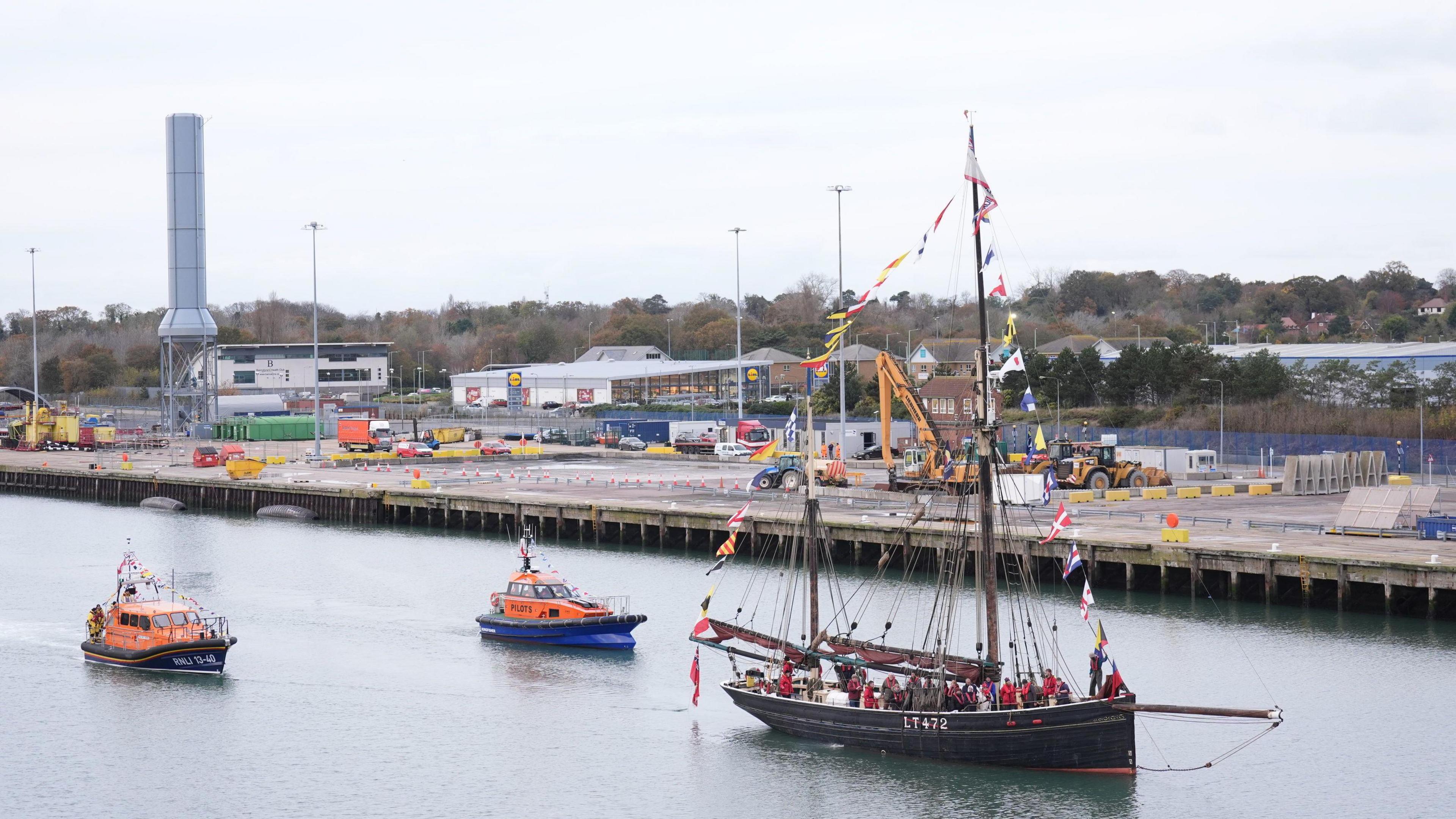 A large traditional fishing boat is pictured sailing on Lake Lothing while it is escorted by two lifesaving boats. The fishing boat has numerous nation flags adorned to its sails.