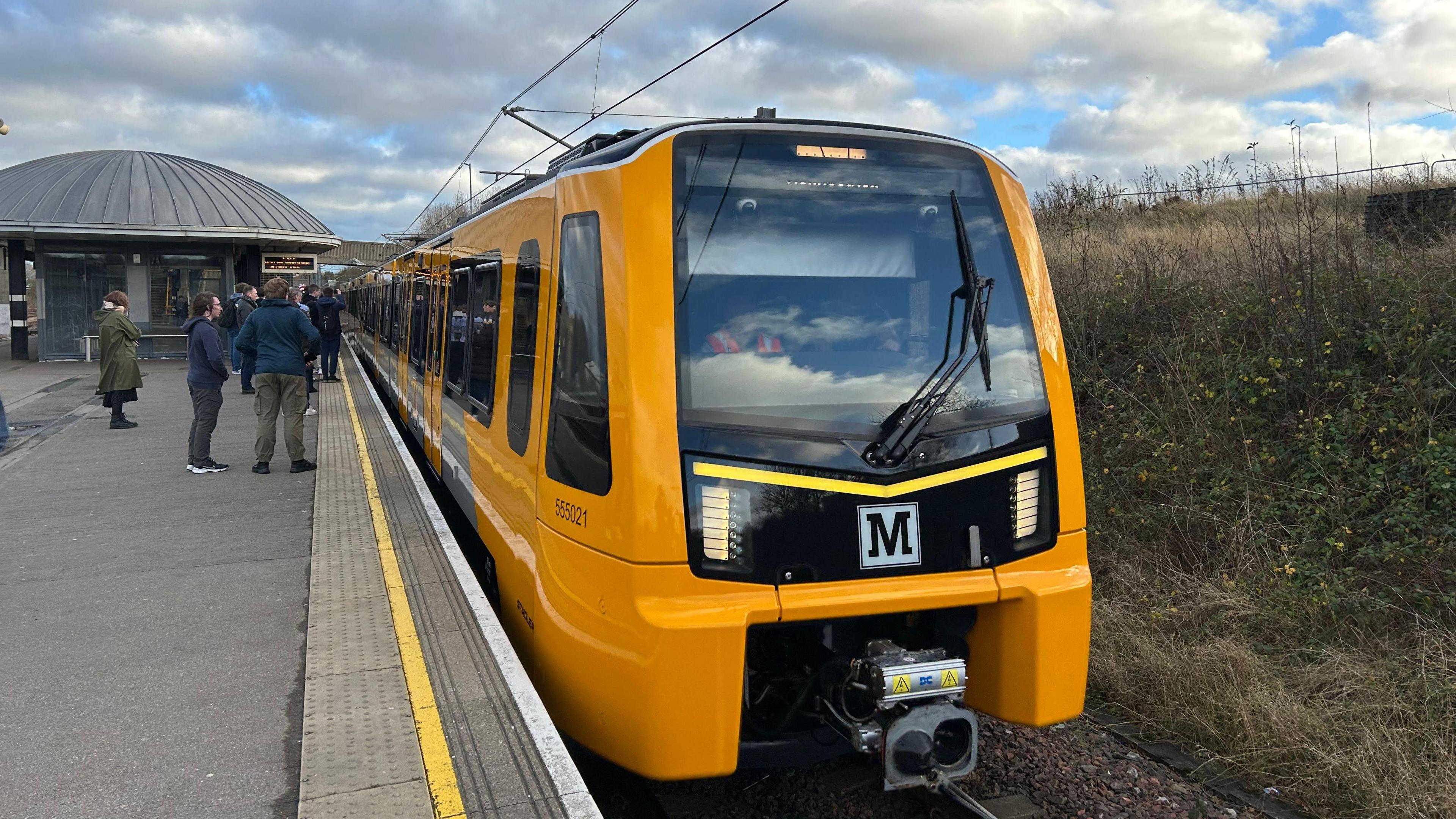 The new Tyne and Wear Metro train which is bright yellow. It's standing at a station and passengers are waiting on the platform.