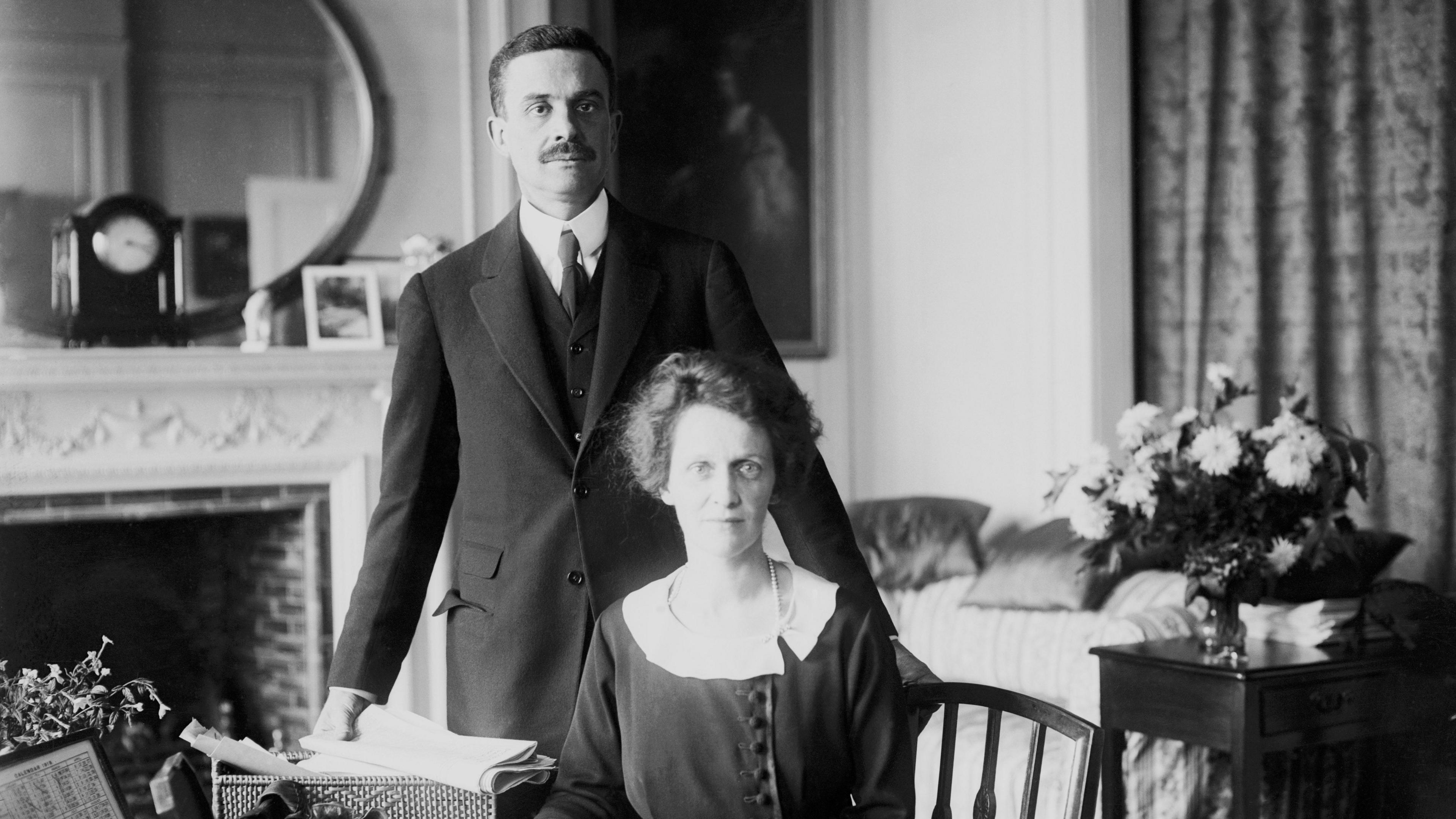 Waldorf Astor, the 2nd Viscount Astor, and his wife Nancy Astor. Nancy is sitting behind a desk and Waldorf is standing up behind her. The photo is black and white and both are wearing period clothing.