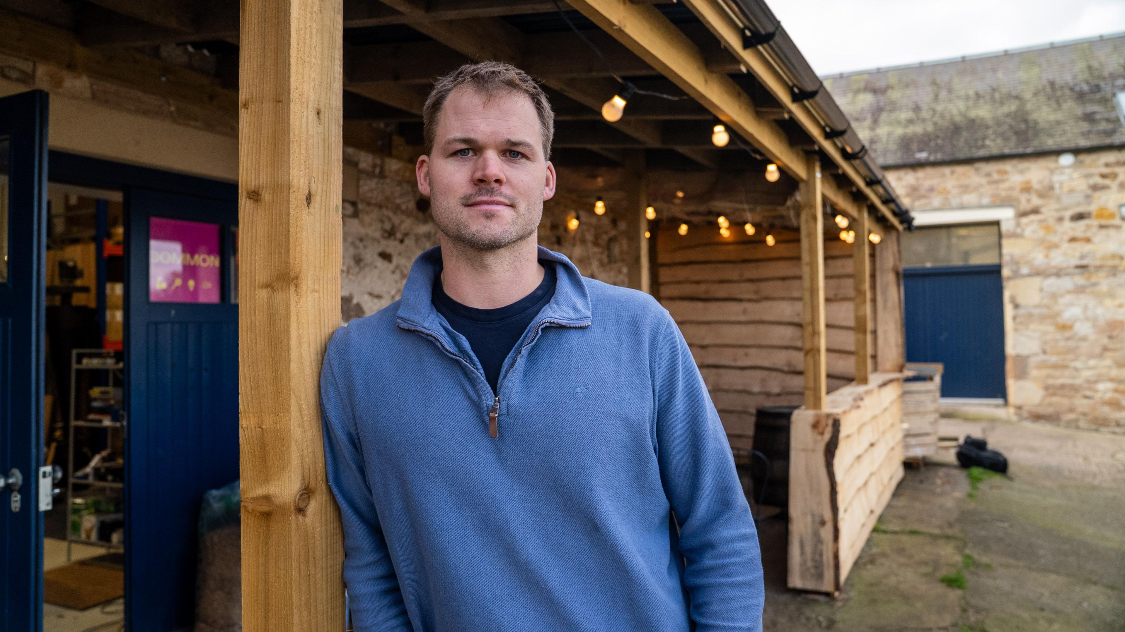 Will Callander looks at the camera as he leans against a wooden post which holds up a shed adorned with fairy lights on the side of a stone building in the courtyard of a farm steading. A dark blue door lies open to the left. Will is wearing a dark blue T-shirt and a light blue V-neck top with a short, open zip. He has brown hair and stubble.
