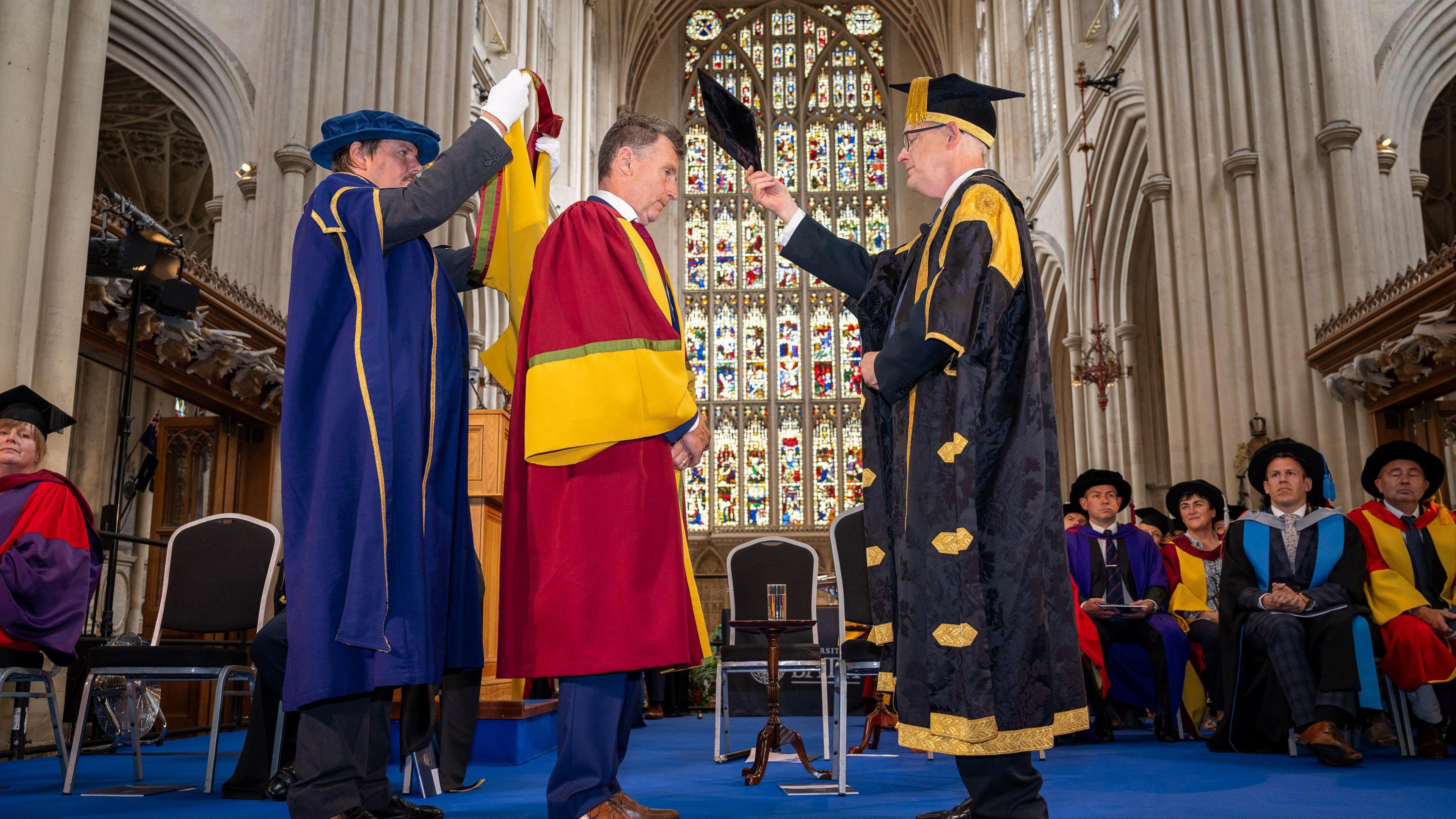 Two men helping placing a graduation cap on another man dressed in red graduation robes