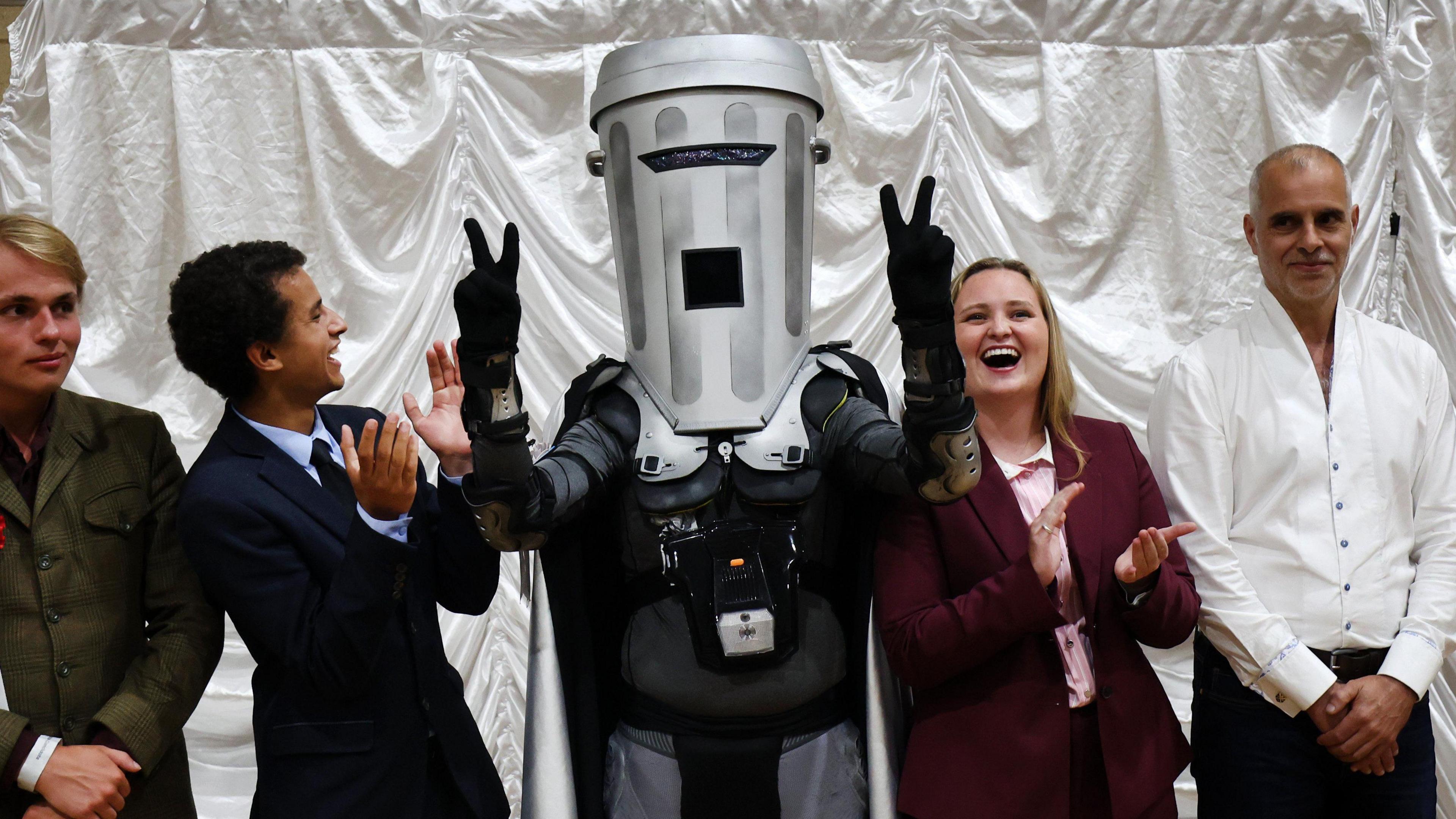 Man wearing grey bin mask over his head lifting two arms and holding his fingers in peace signs