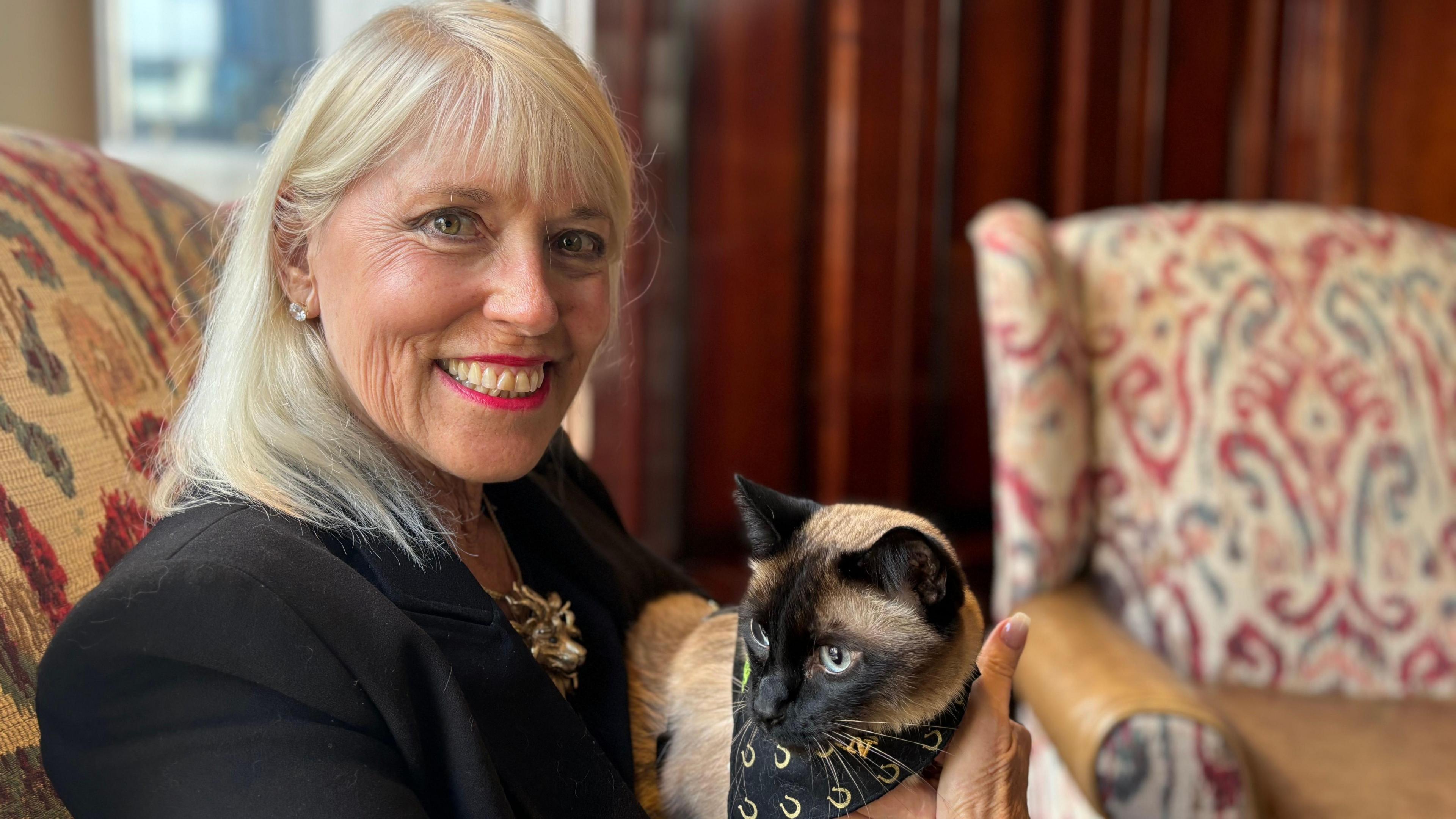 Blonde woman in all black smiles as she holds her Siamese cat on her lap. 