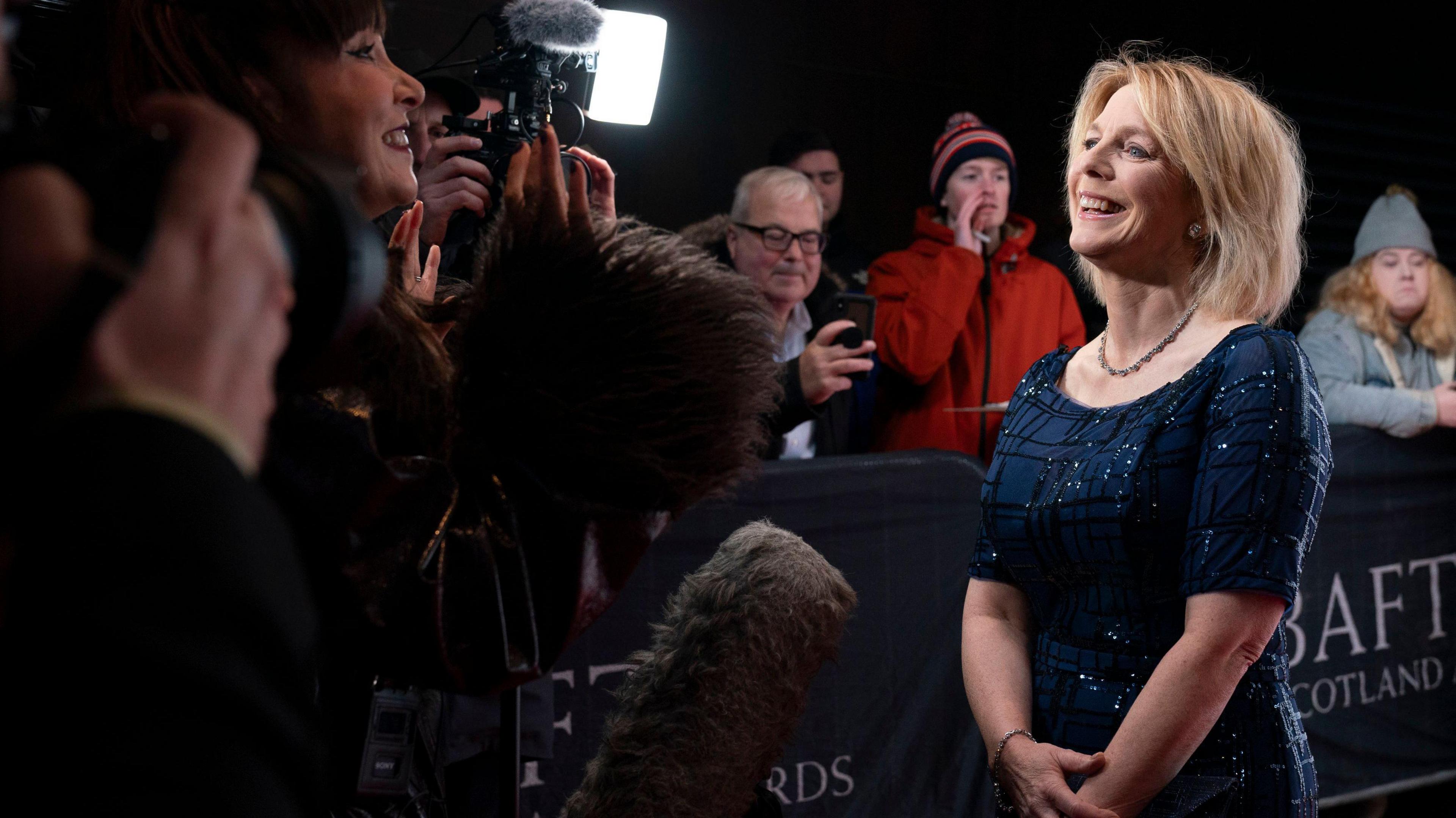 Hazel Irvine arriving at the BAFTA Scotland Awards, facing reporters and journalists. There are members of the public behind a barrier. She is wearing a navy blue dress with sequins sparkling in the lights.