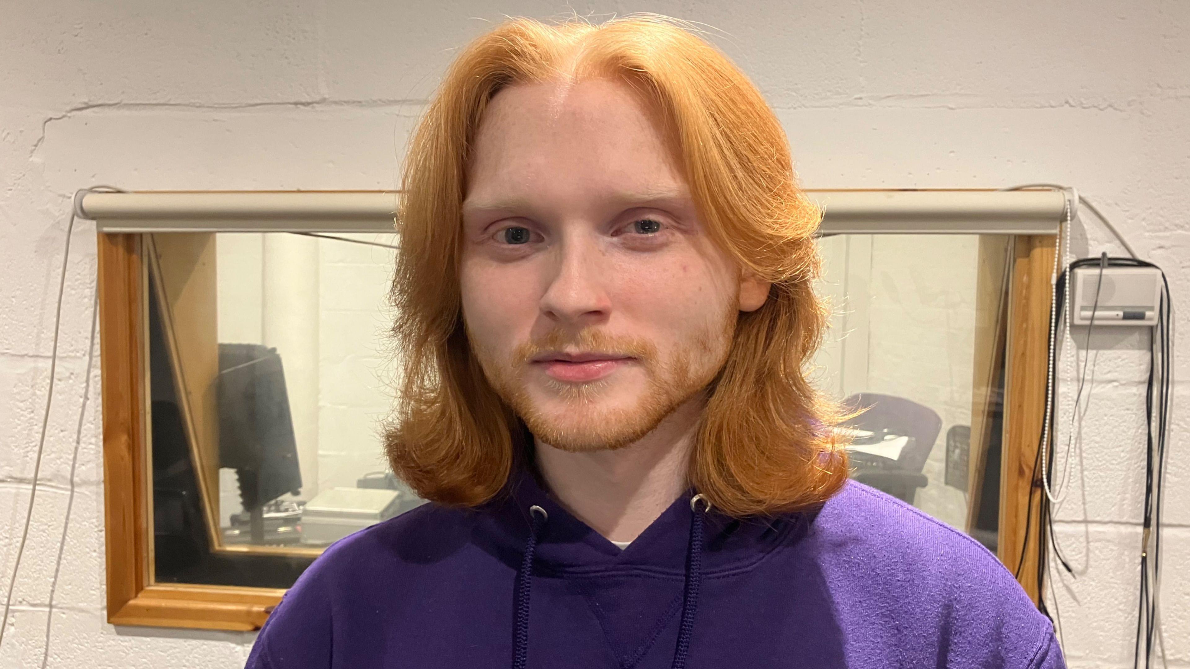 Harry looks into the camera in a head and shoulders image. He is wearing a purple hoodie and has long ginger hair down to his shoulders, and a cropped ginger beard.