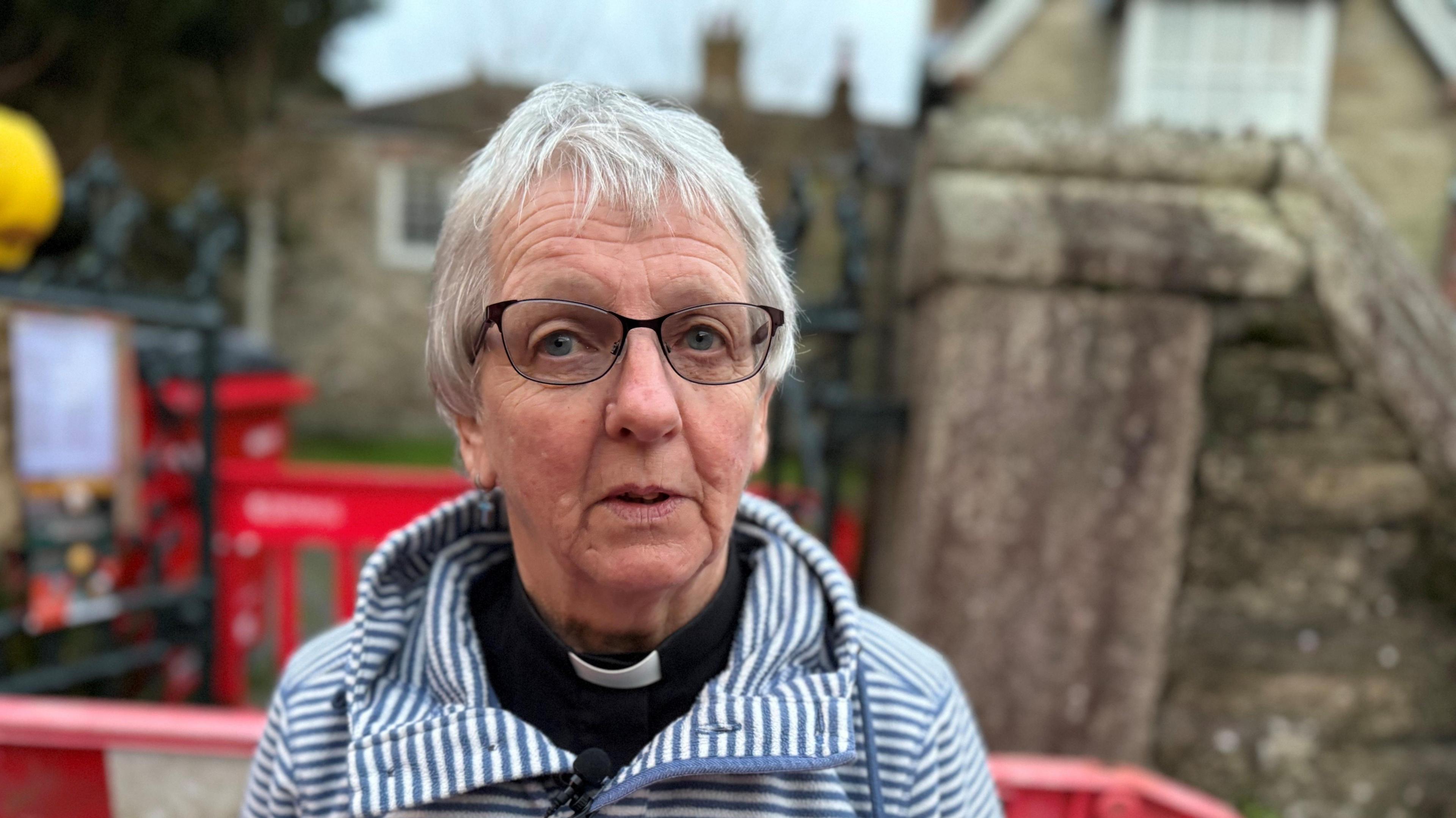 Older person with white hair and glasses and wearing a dog collar and fleece on top looking at camera