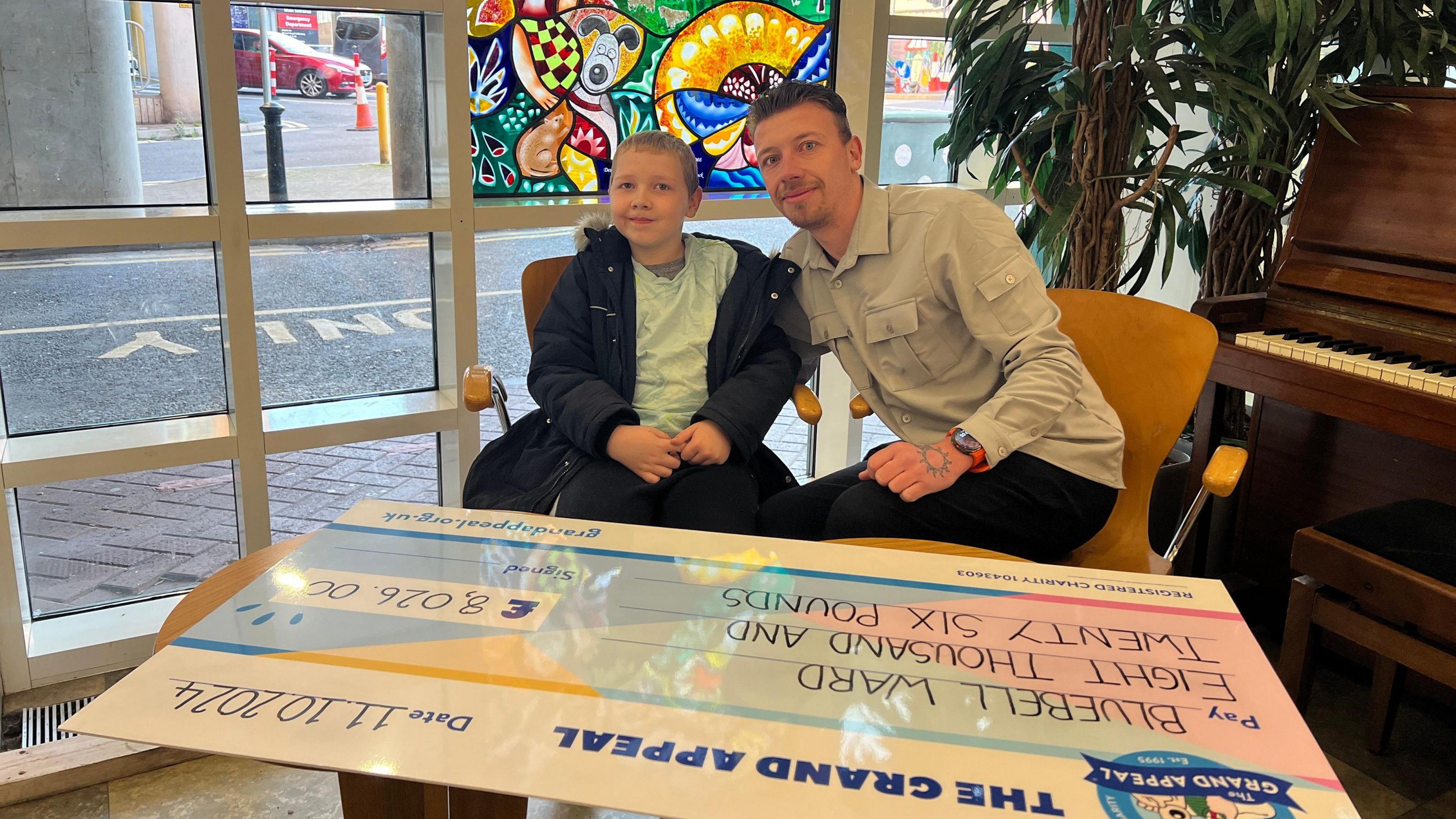 Harry (left) and father (Edward) seen smiling at the camera. They are sitting in front of the giant cheque, which they are about to sign. 