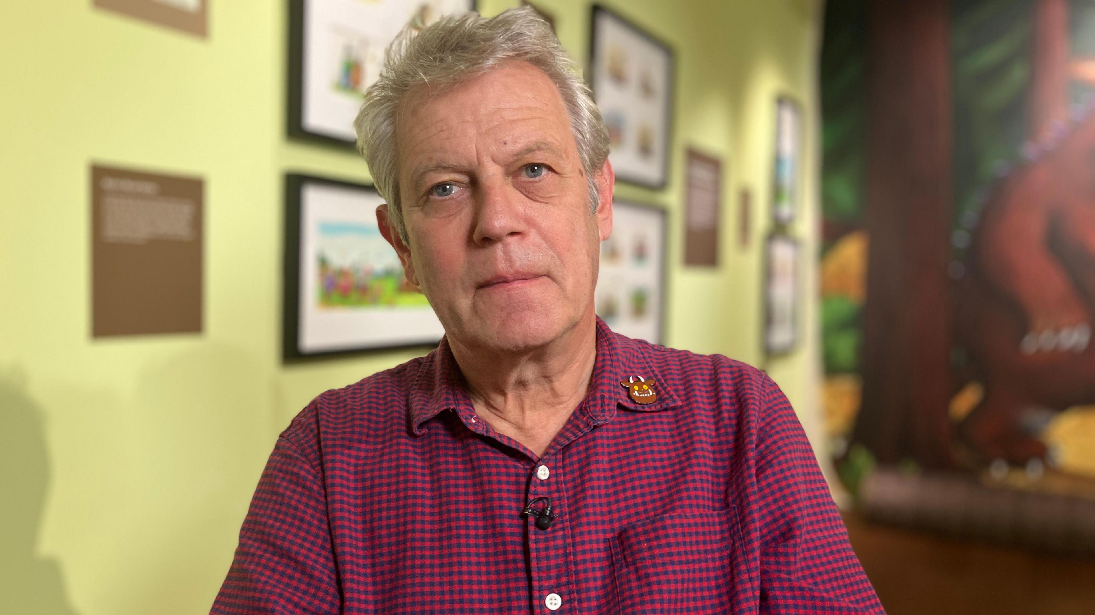 Axel Scheffler is wearing a burgundy checked shirt. He sits in front of some of his framed illustrations