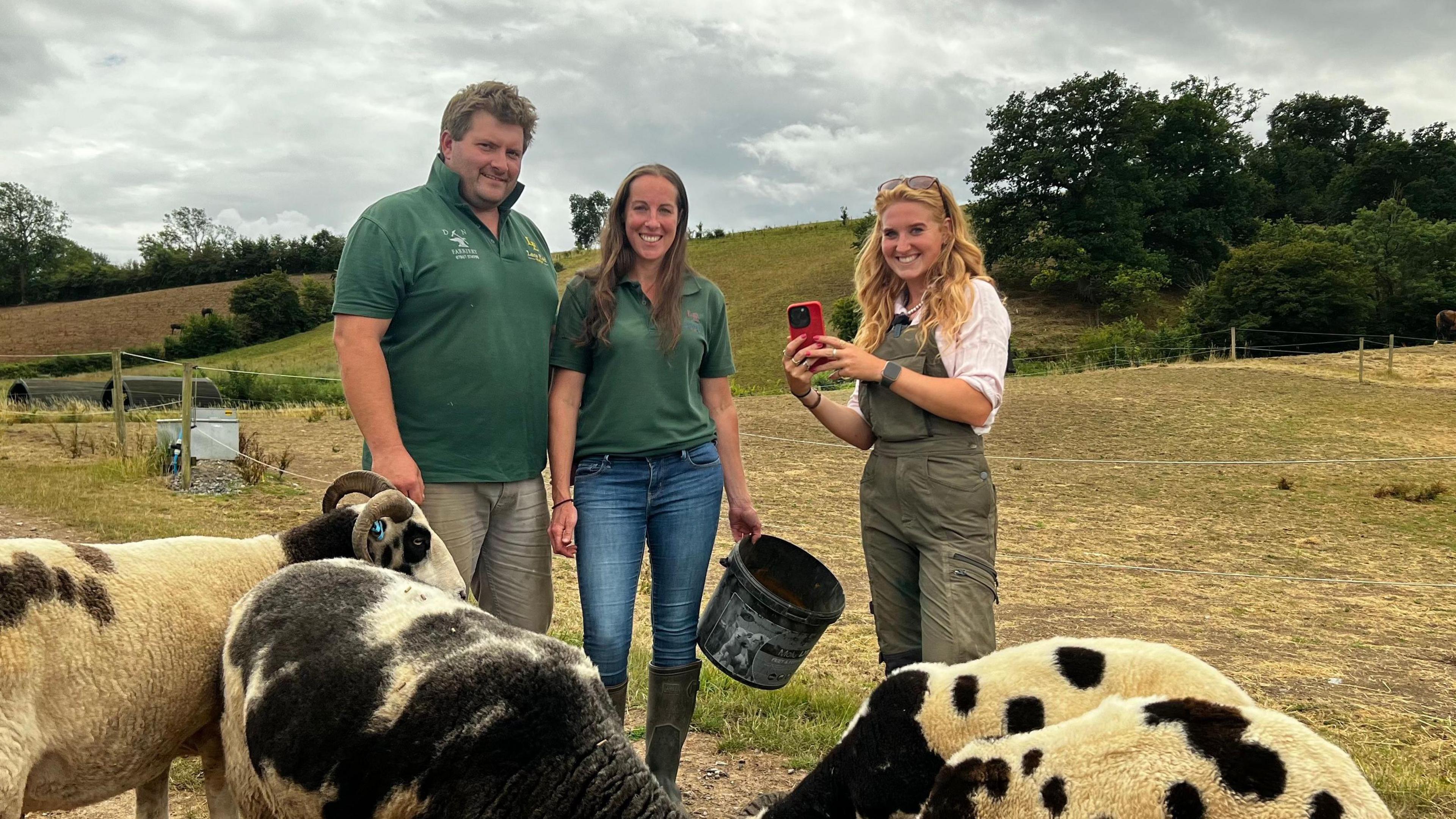 A man and two women stand in a field with black and white sheep grazing in the foreground. To the left a man looks at the camera. he has tousled light brown hair and is wearing a bottle green coloured short sleeve collared shirt. A woman wearing a similar shirt and blue jeans stands next to him smiling at the camera and holding a black feed bucket in one hand. Next to her is Chloe Burke who is holding a red mobile phone up as if she is filming, while smiling.