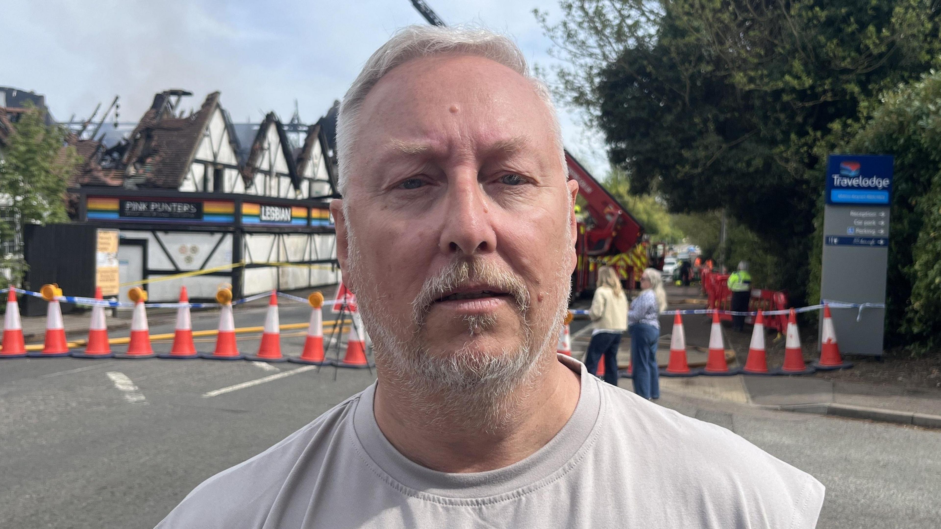 A man with grey hair and a grey beard looks at the camera as he stands on a closed road in front of a nightclub where a fire has been. He wears an off-white top. People can be seen looking at the venue behind him. There is a cordon of cones in front of the venue.