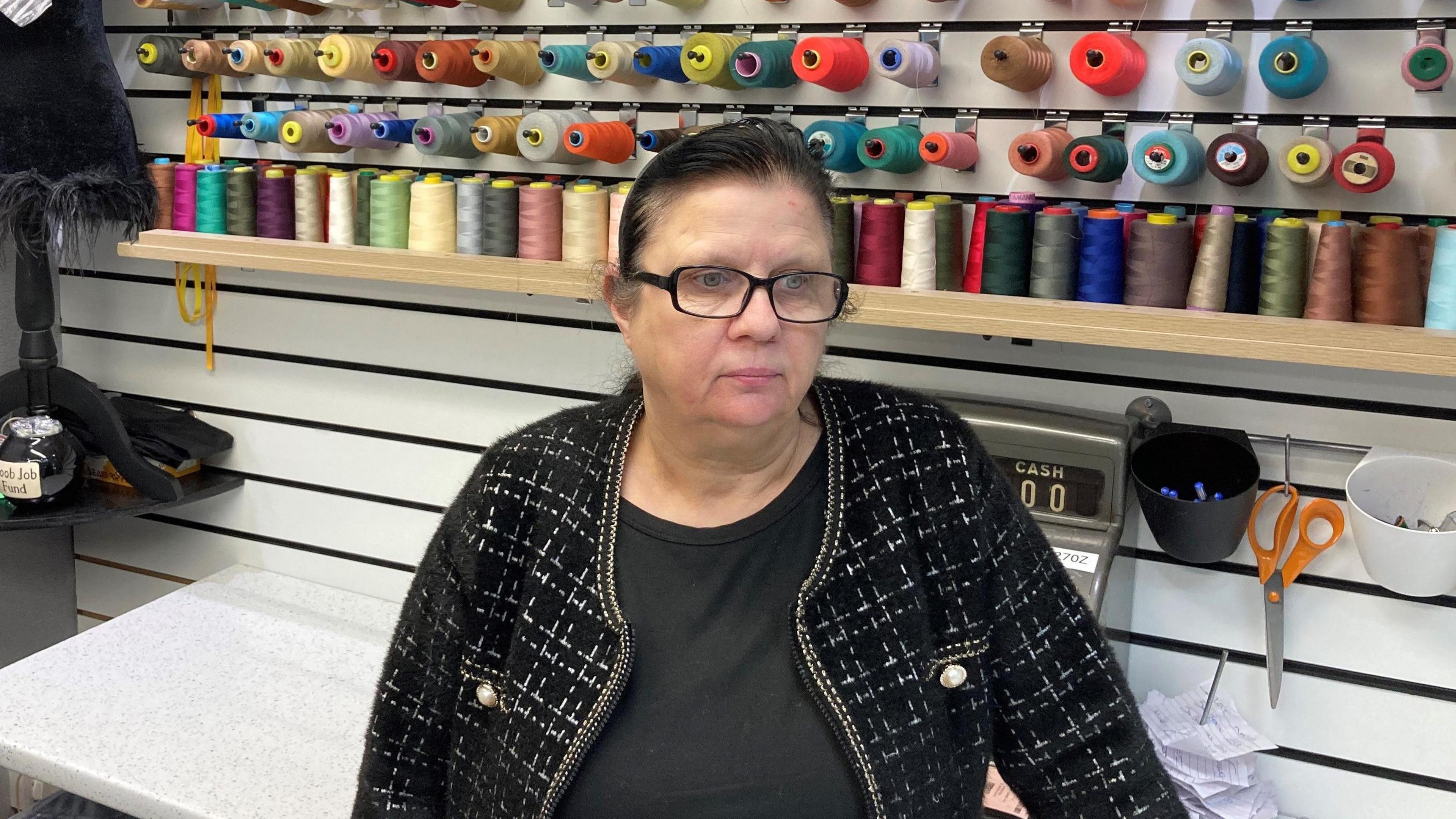 Boowy Latham, who has black hair and black framed glasses, speaks to the camera in front of a shelf holding spools of coloured thread.