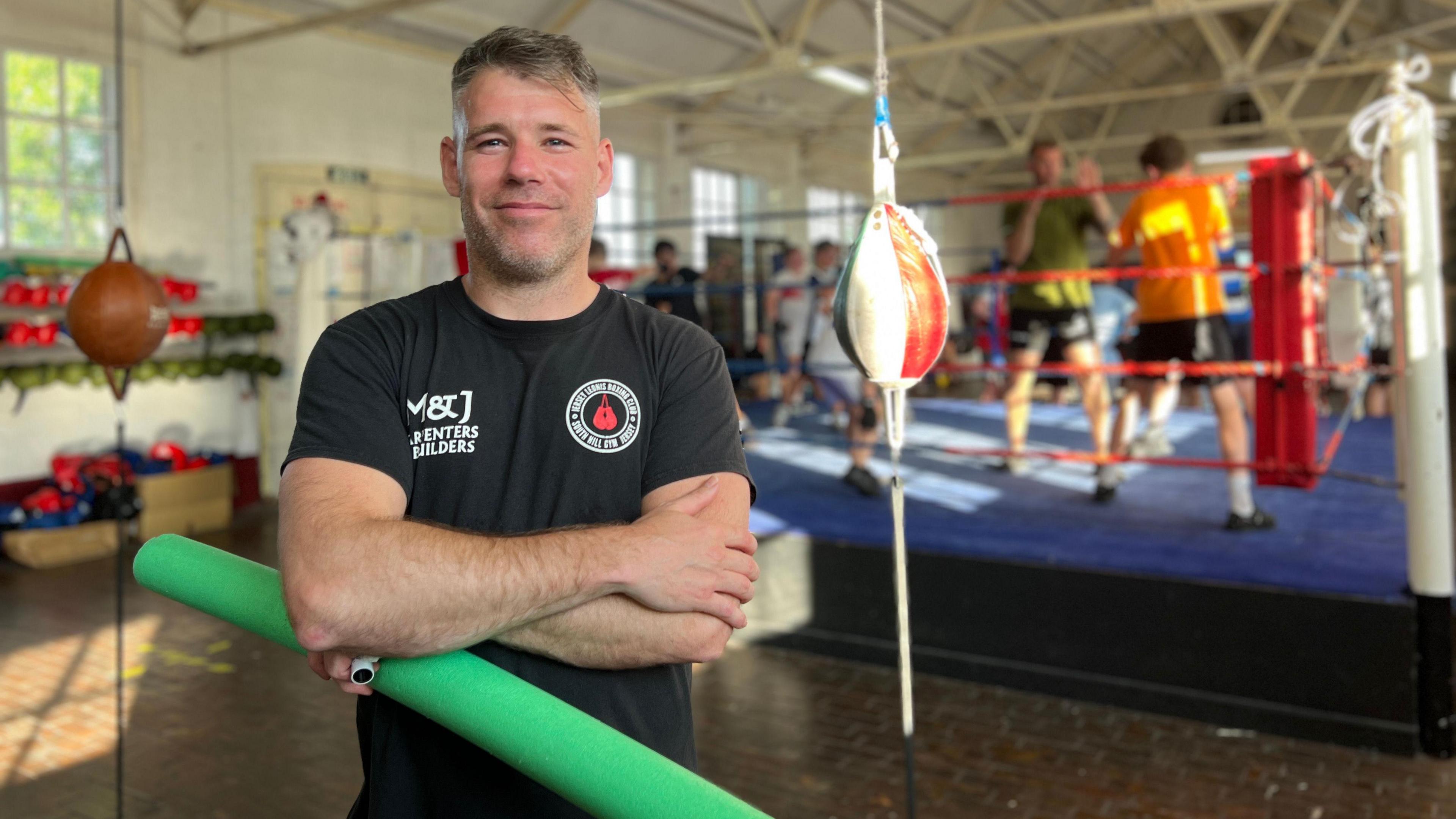 Harry stands in the foreground of a boxing gym, holding a green foam roller. He wears a black t-shirt with white text and logos. Behind him, several people are training in a boxing ring. The gym features high ceilings with exposed beams, large windows, and various boxing equipment including punching bags and gloves.