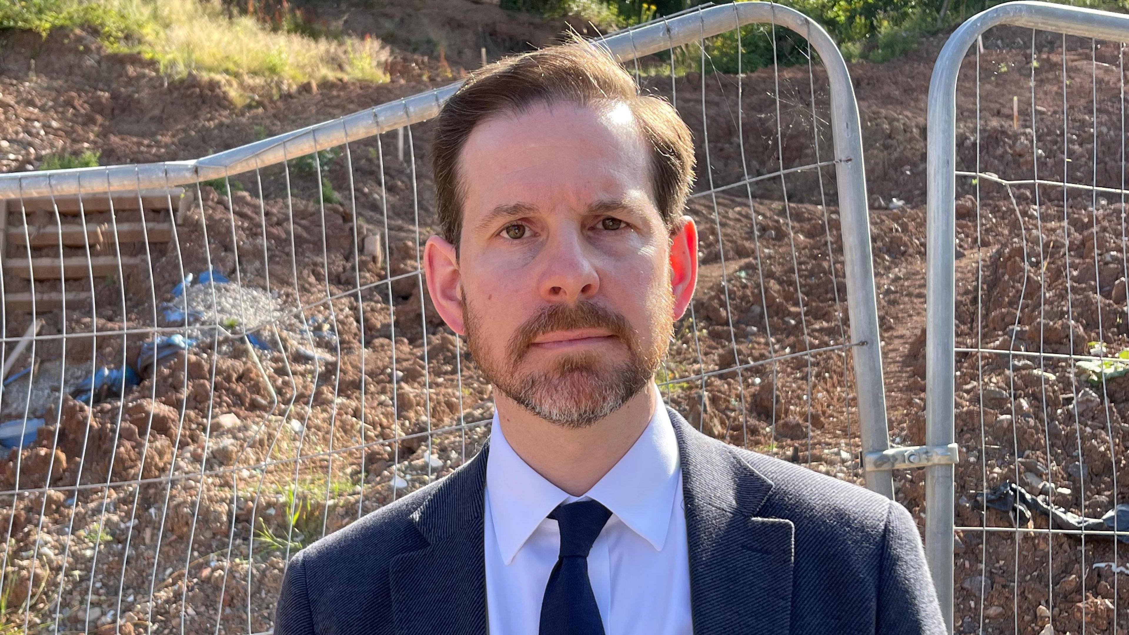 Alex Ballinger MP standing in front of the landslip. He is wearing a black tie, white shirt and dark blue blazer. He has a short dark beard and short dark hair.