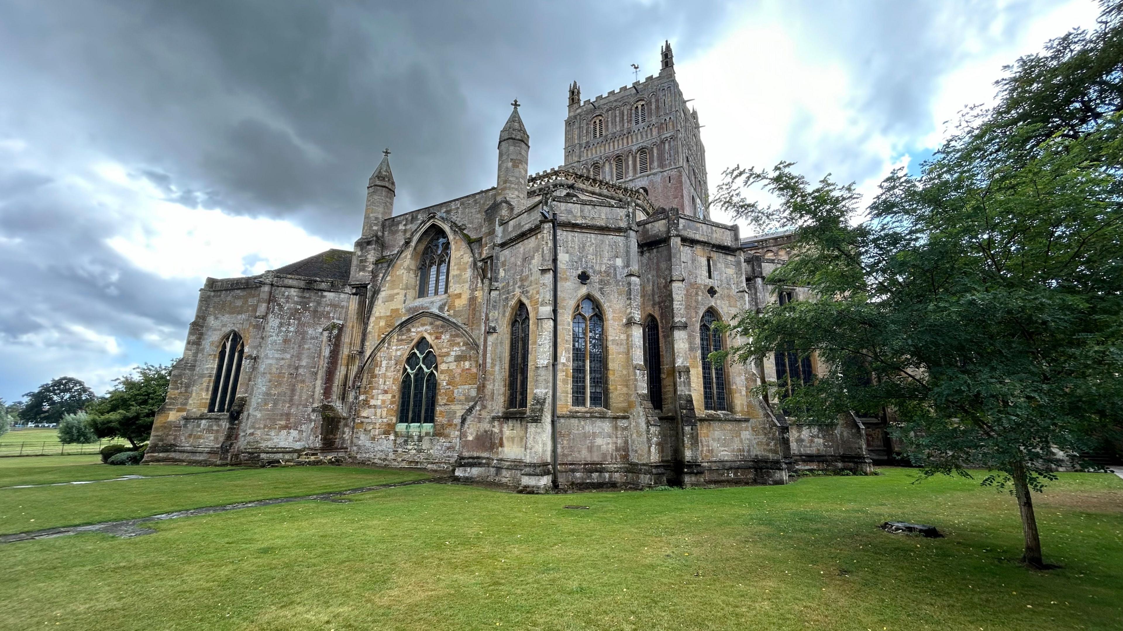 Tewkesbury Abbey, a large church, stands on a very green, cut lawn which has a couple of trees on it. There is a grey, cloudy sky above it with a few patches of sunlight illuminating some clouds.