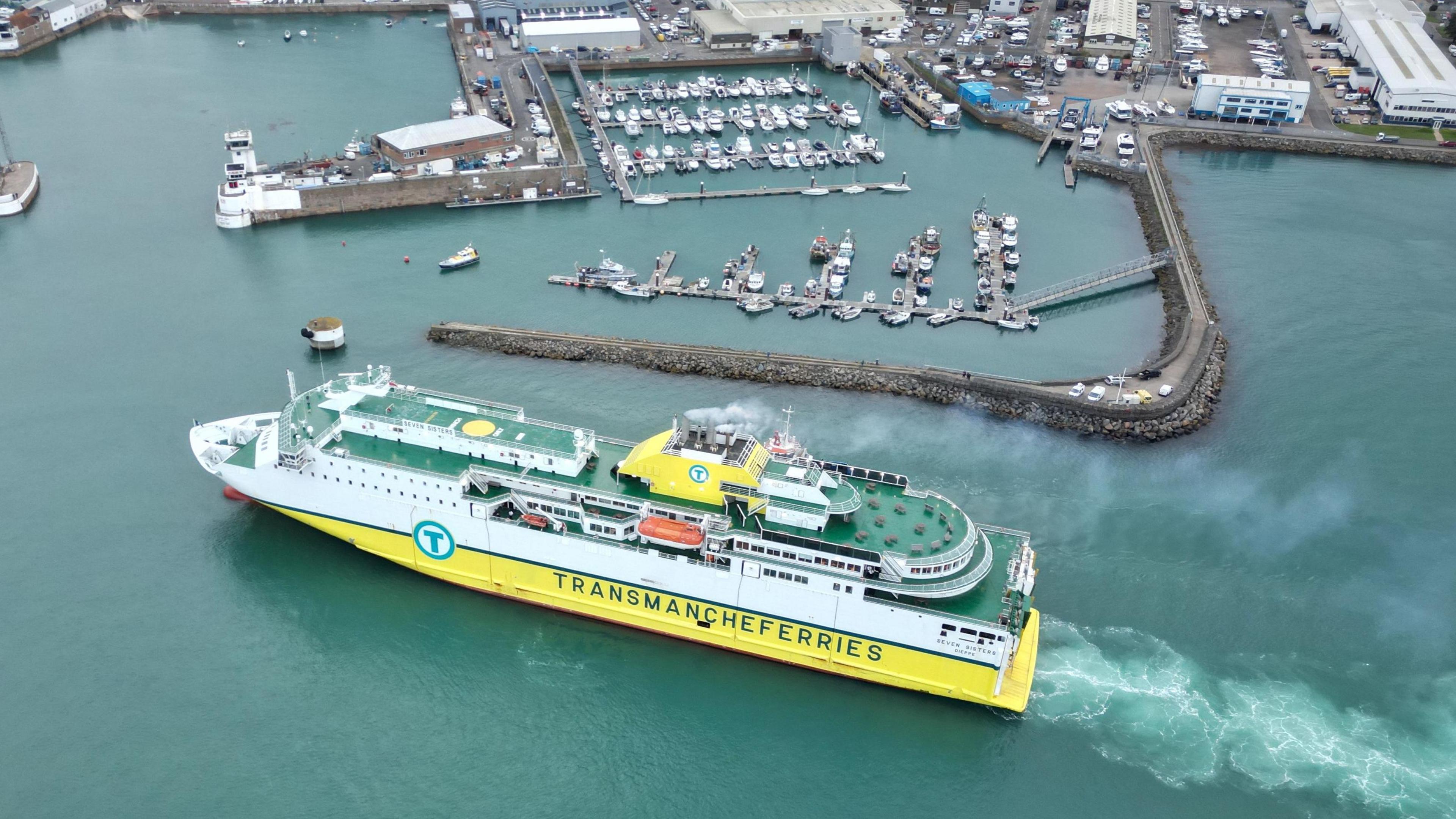 DFDS' Seven Sisters ship coming into Jersey Harbour. The picture was taken from a drone. It shows a large white and yellow ship with green decks. The docks of the harbour are visible behind the ship. With smaller vessels showing the scale of Seven Sisters 