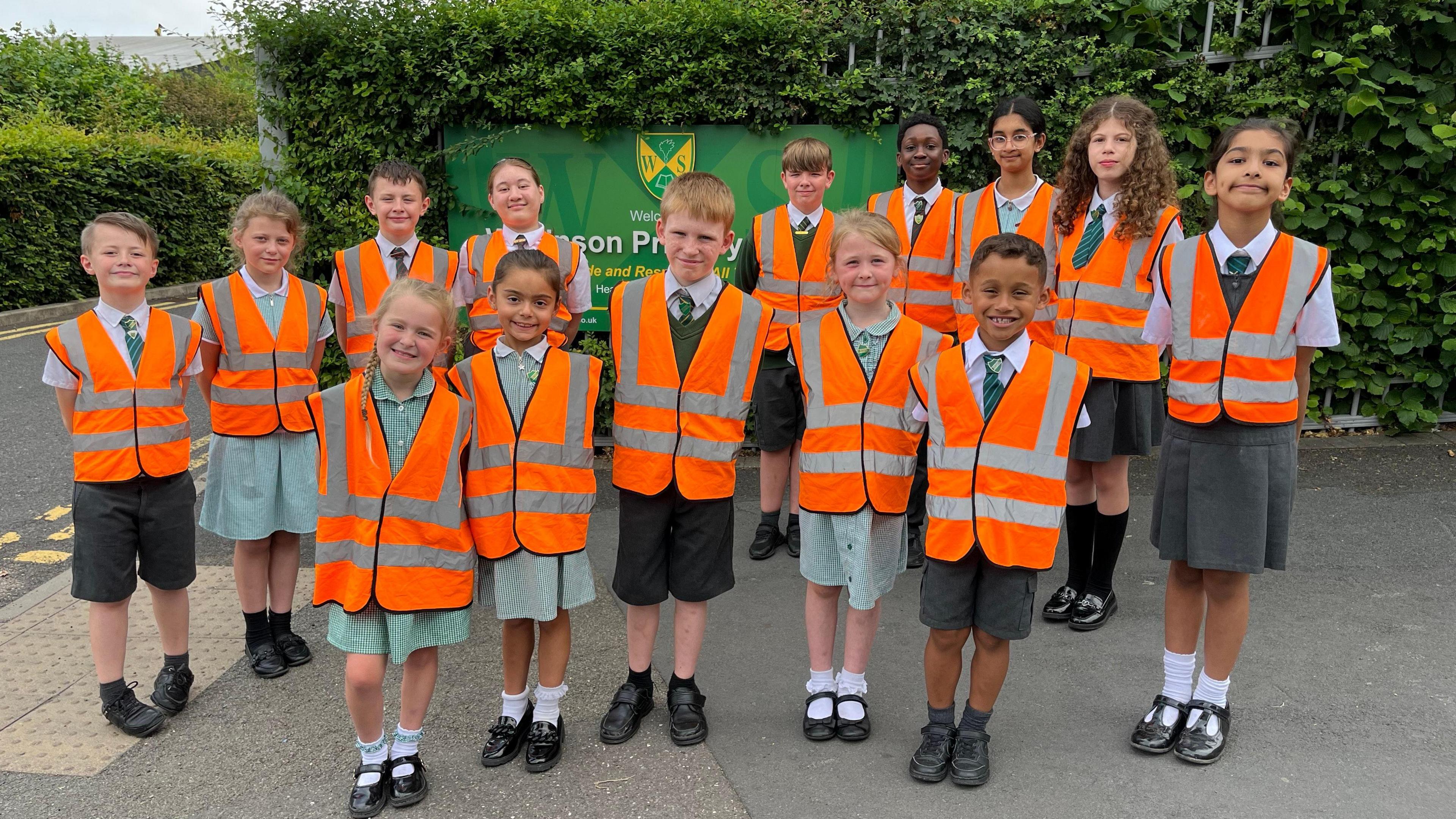 Fourteen pupils, with orange bibs over their uniforms, stand outside the school gates. The school sign can be seen behind them.