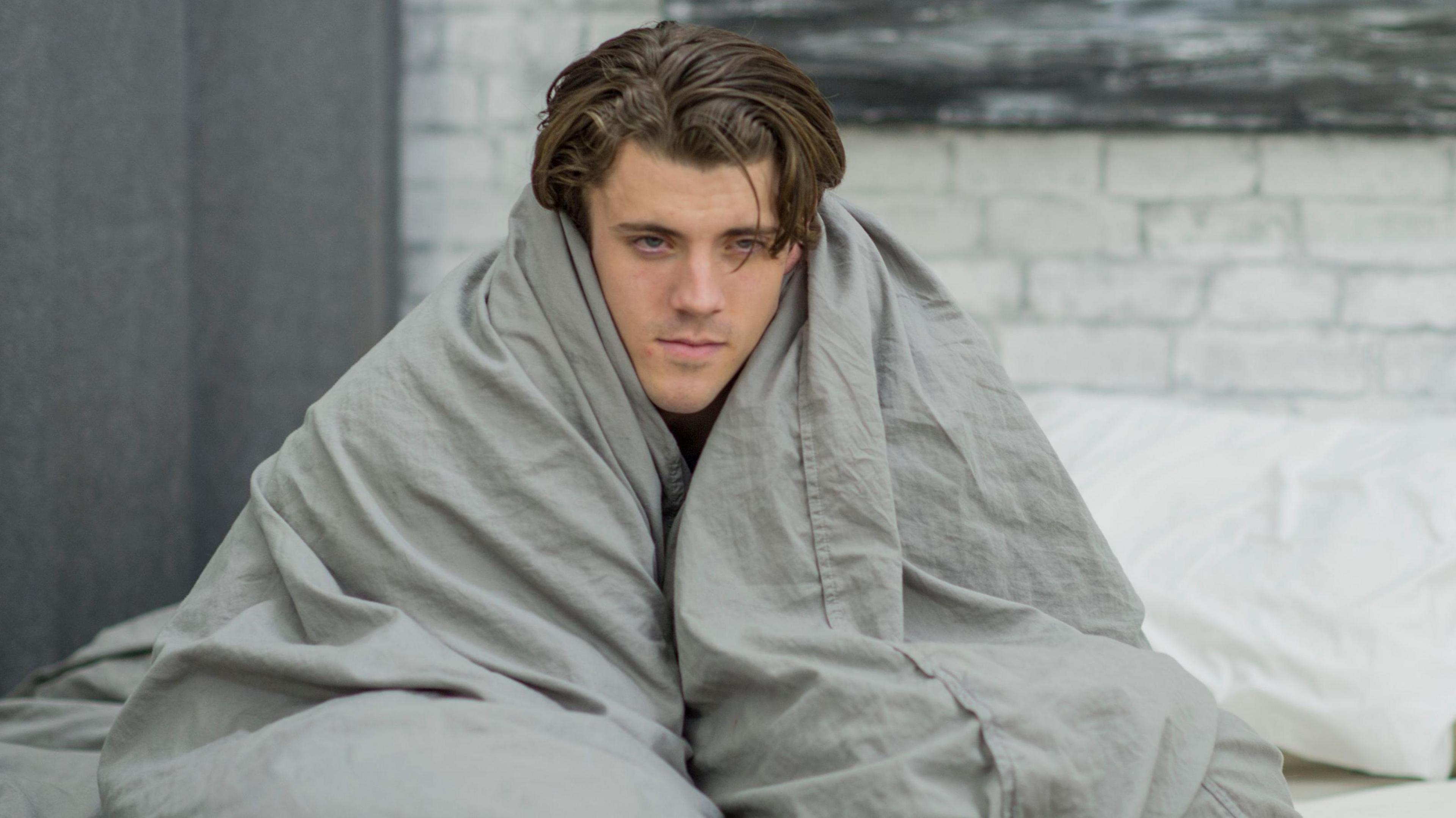 A young man with tousled brown hair is looking glum and a bit ill, wrapped up in a grey-coloured duvet while sitting on his bed