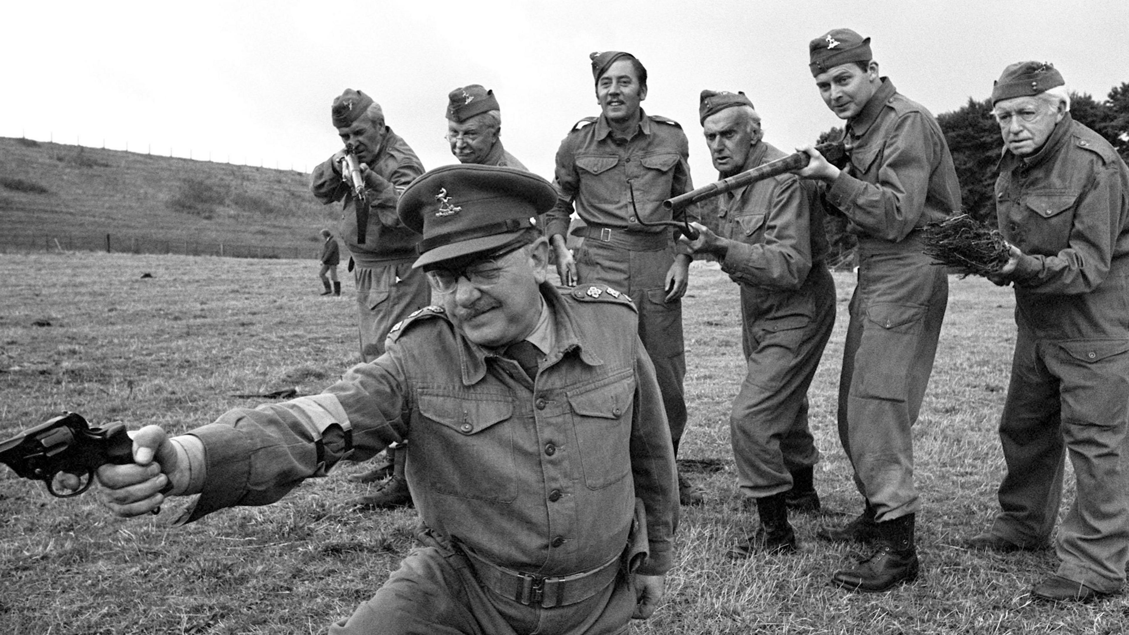 Black and white image dated 20/08/70 of the cast from the BBC's hit comedy Arthur Lowe (foreground) and (back, left to right) John Le Mesurier, Clive Dunn, James Beck, John Laurie, Ian Lavender and Arnold Ridley, in a scene from one of the famous episodes of the show. They are wearing Army uniforms and are outside in a grassy field. Arthur Lowe is kneeling with his right arm outstretched holding a pistol. The others are lined up behind him holding rifles. 