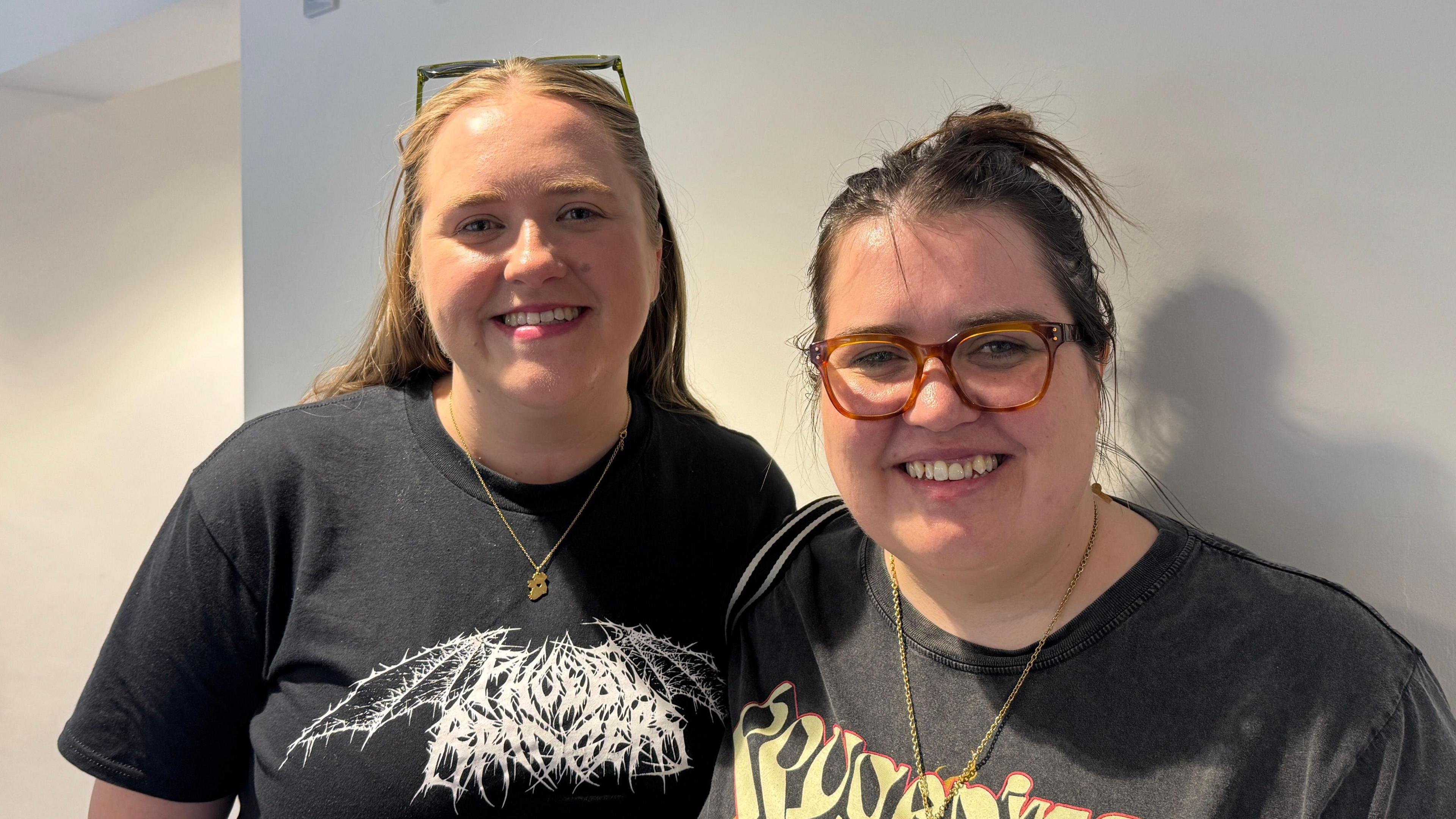 Sisters Eibhlin and Emma smile at the camera while they have their arms around each other. Eibhlin on the left is wearing a black Phoebe Bridgers t-shirt, a gold necklace and has long, blonde hair. Emma, on the right, has her brown hair tied back and is wearing a pair of brown glasses.