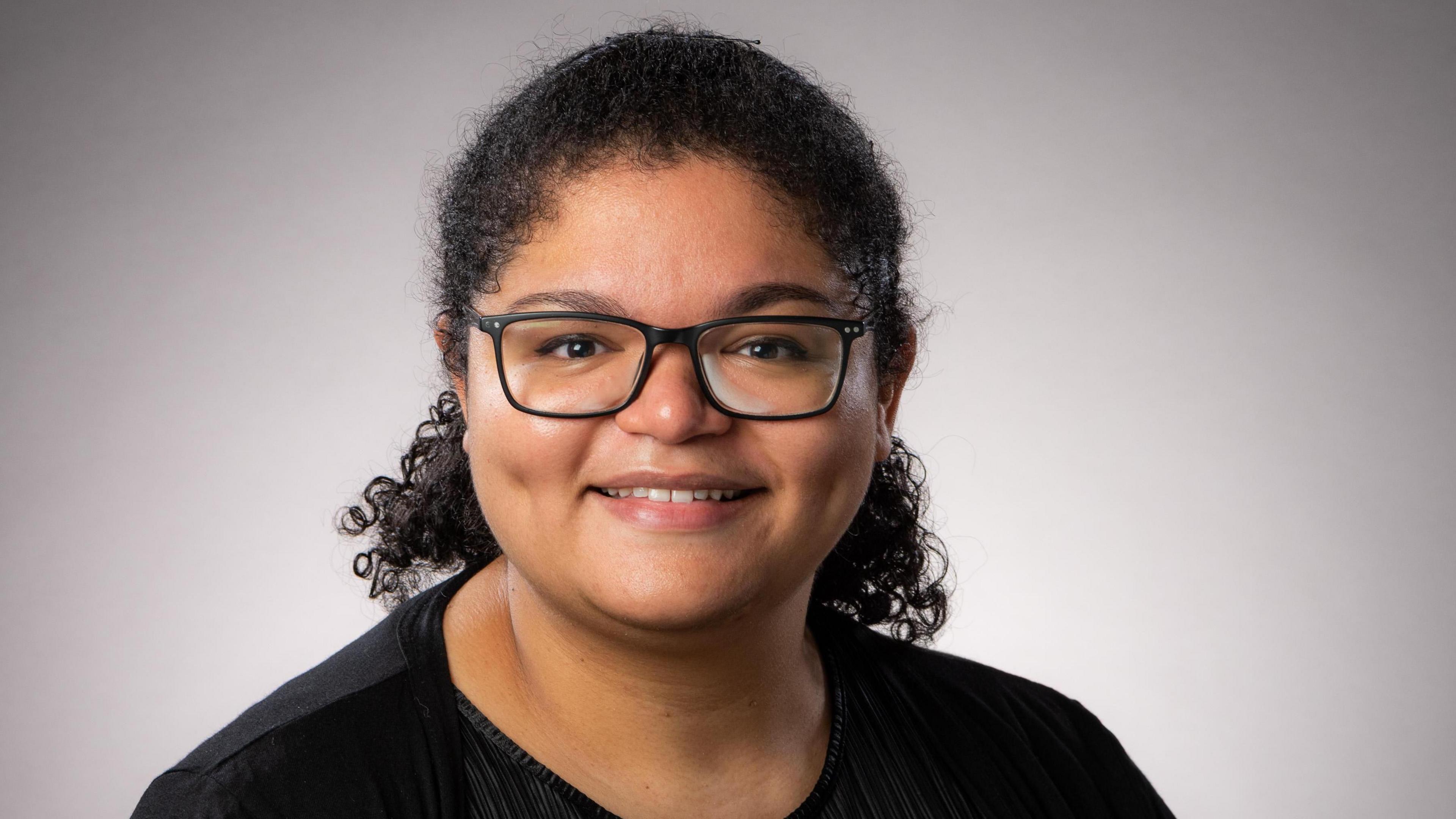 A professional headshot of Dr Nina Higson-Sweeney wearing a black t shirt and black rectanglar framed glasses 