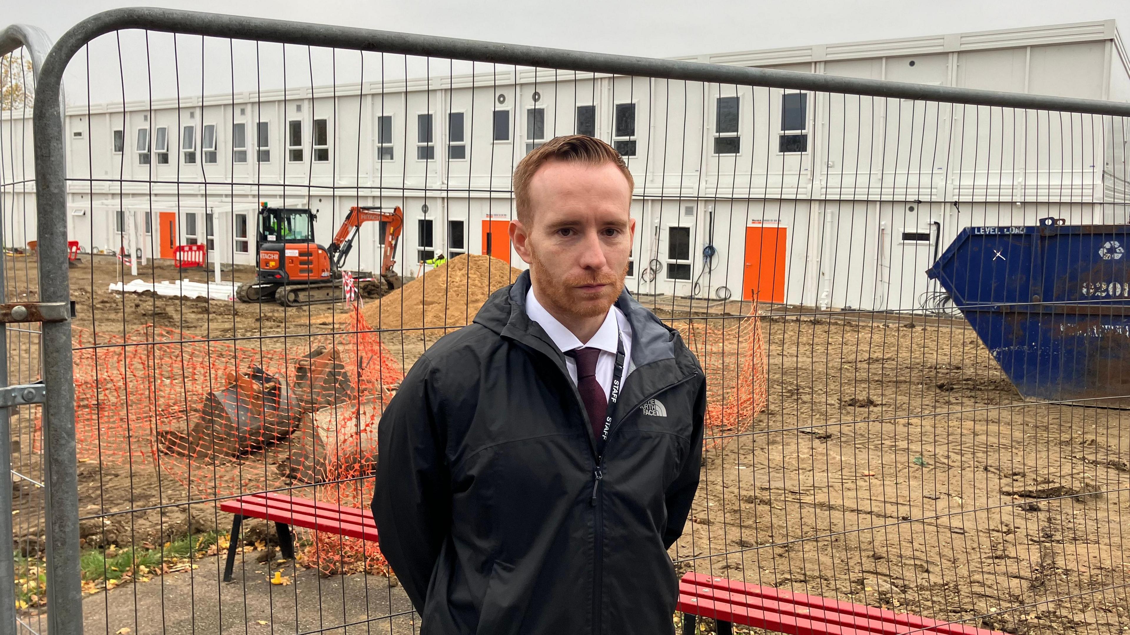 A head-and-shoulders picture of Katherines Head of School, Julien Mealey. He has short, red hair and a beard. He is wearing a black coat and a white shirt and black tie. He is standing in front of a fence. On the other side of the fence, there is a building site. A two-storey temporary building is being constructed.