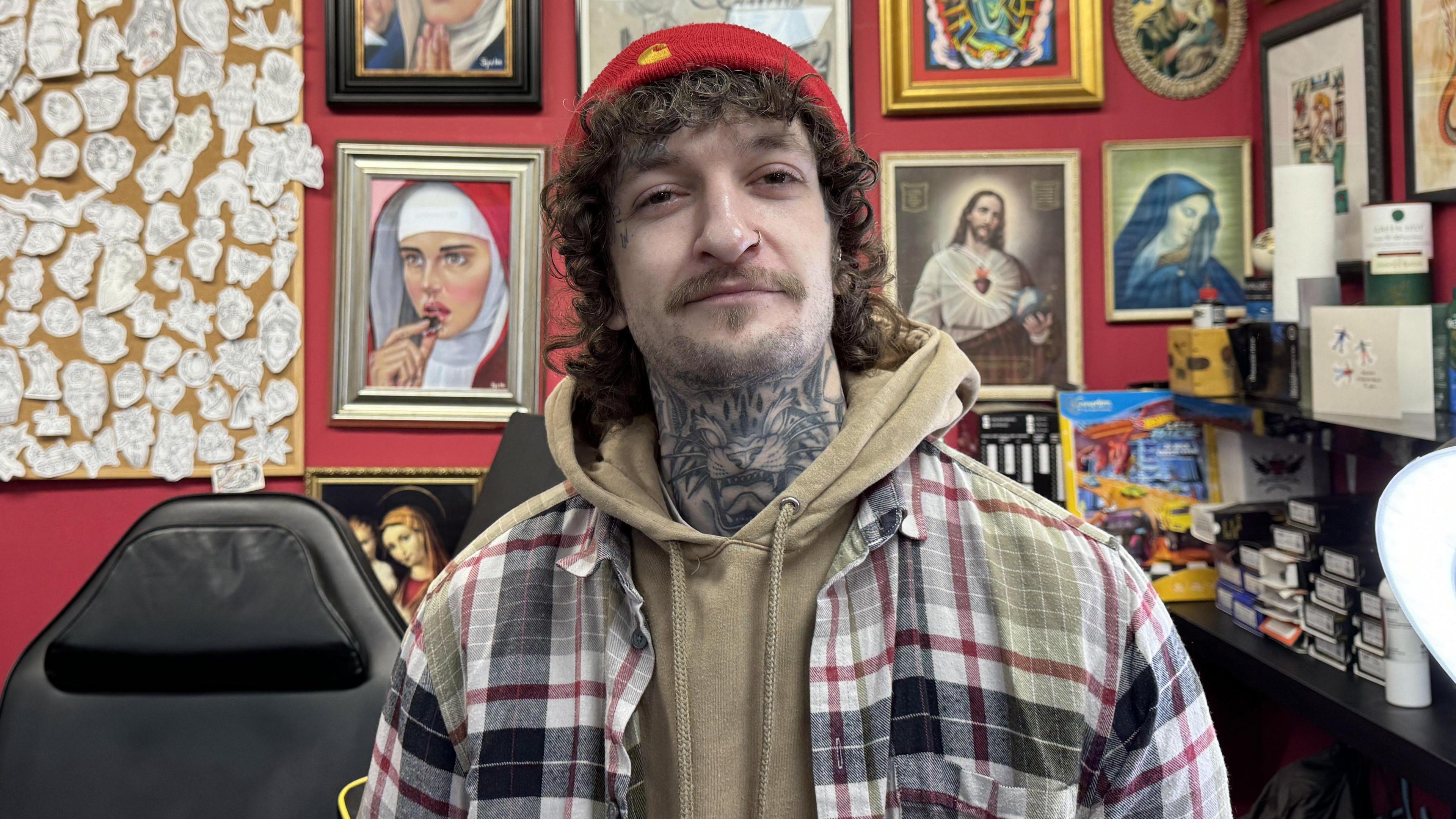 Ross Walker, he is wearing a red hat, light brown jumper and check shirt standing in his tattoo parlour. 