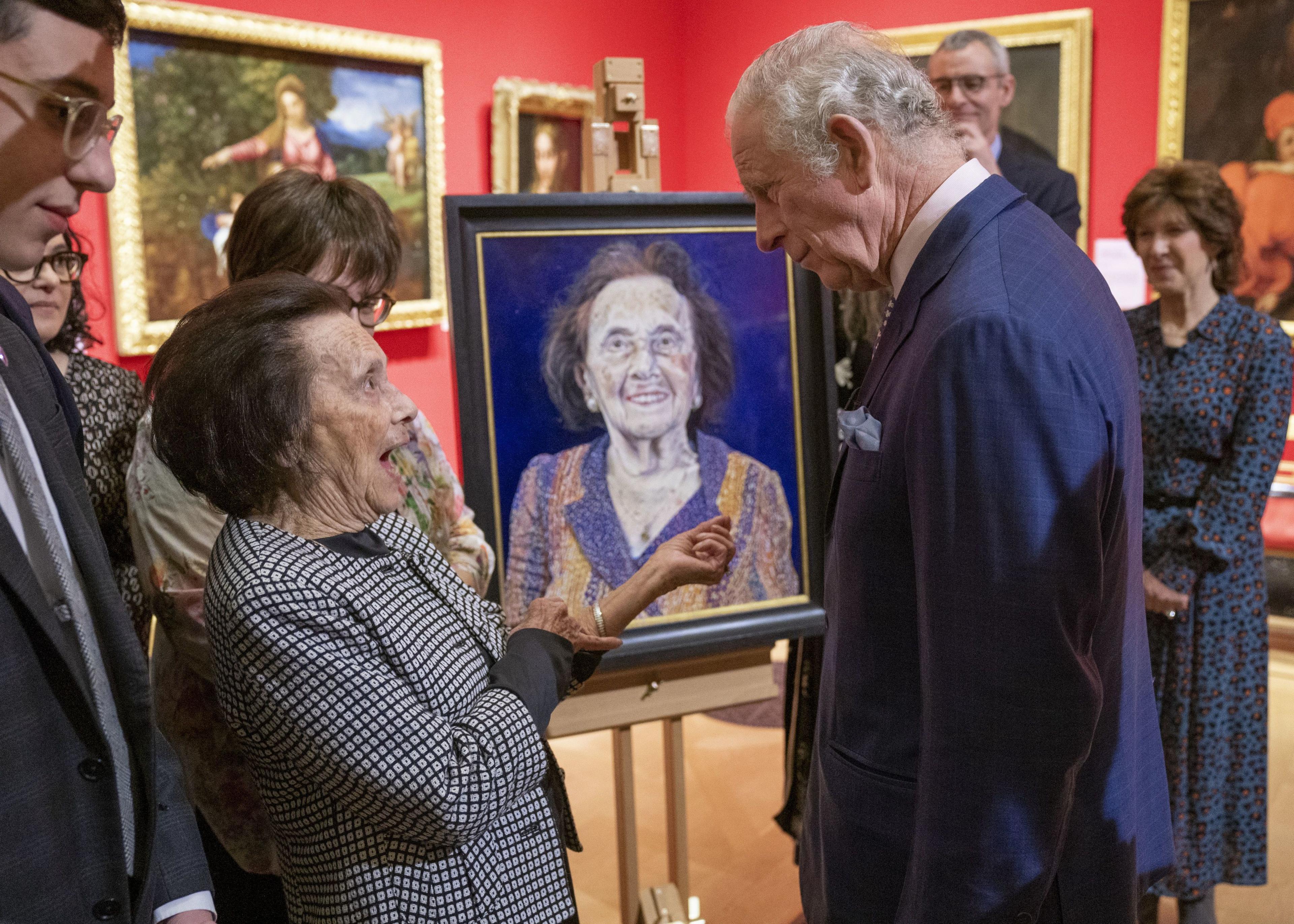 Lily, wearing a black and white blazer, and King Charles, in a navy suit, having a conversation in front of her portrait.