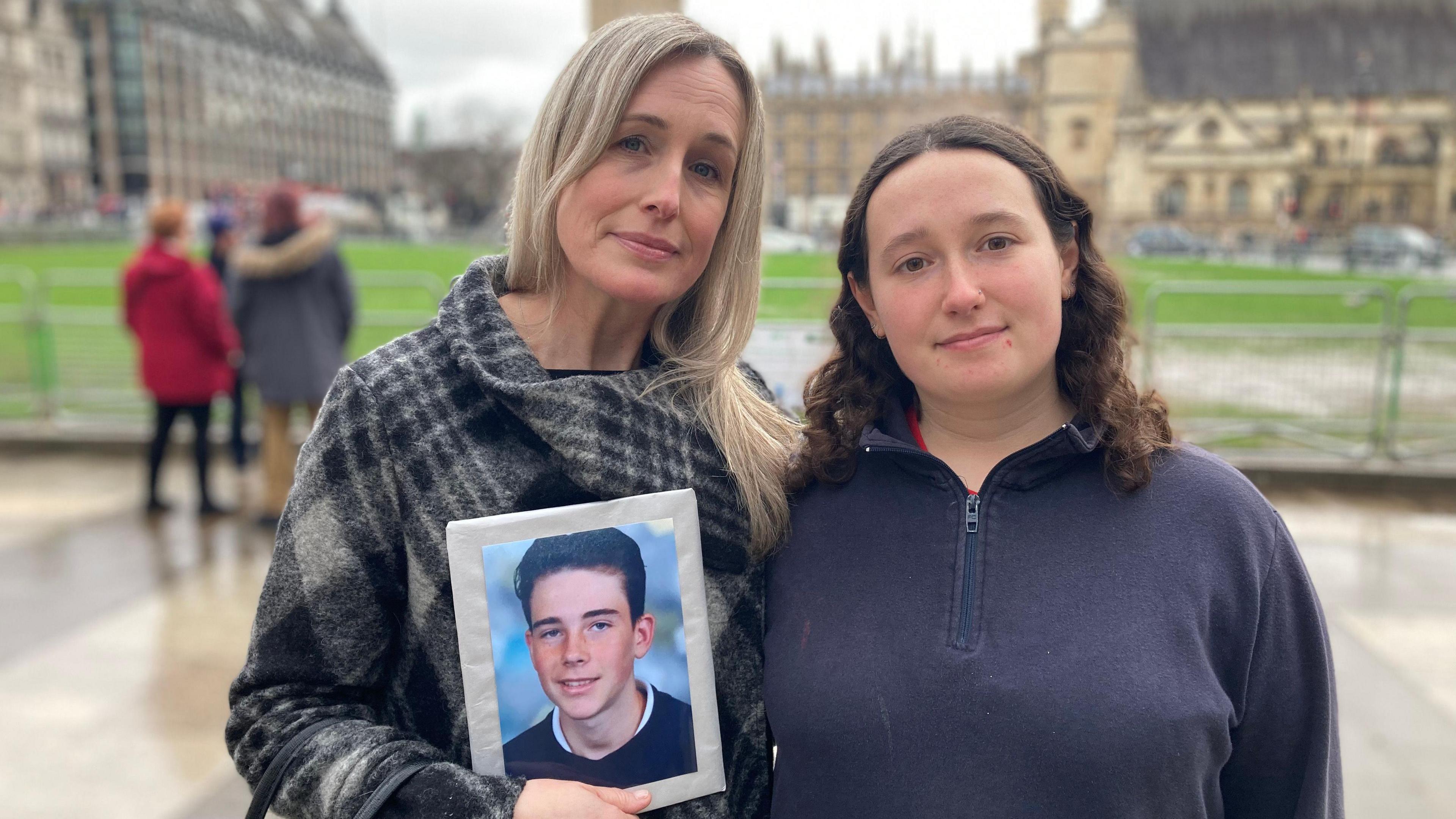 Sam Robinson and Mia Pullen pose with Parliament Square in the background. Sam has long blond hair, and is wearing a black and white checked coat. She is holding up a photo of her son Billy. Next to her is Mia Pullen, wearing a dark blue quarter zip jumper. She has shoulder length brown, wavy hair. 