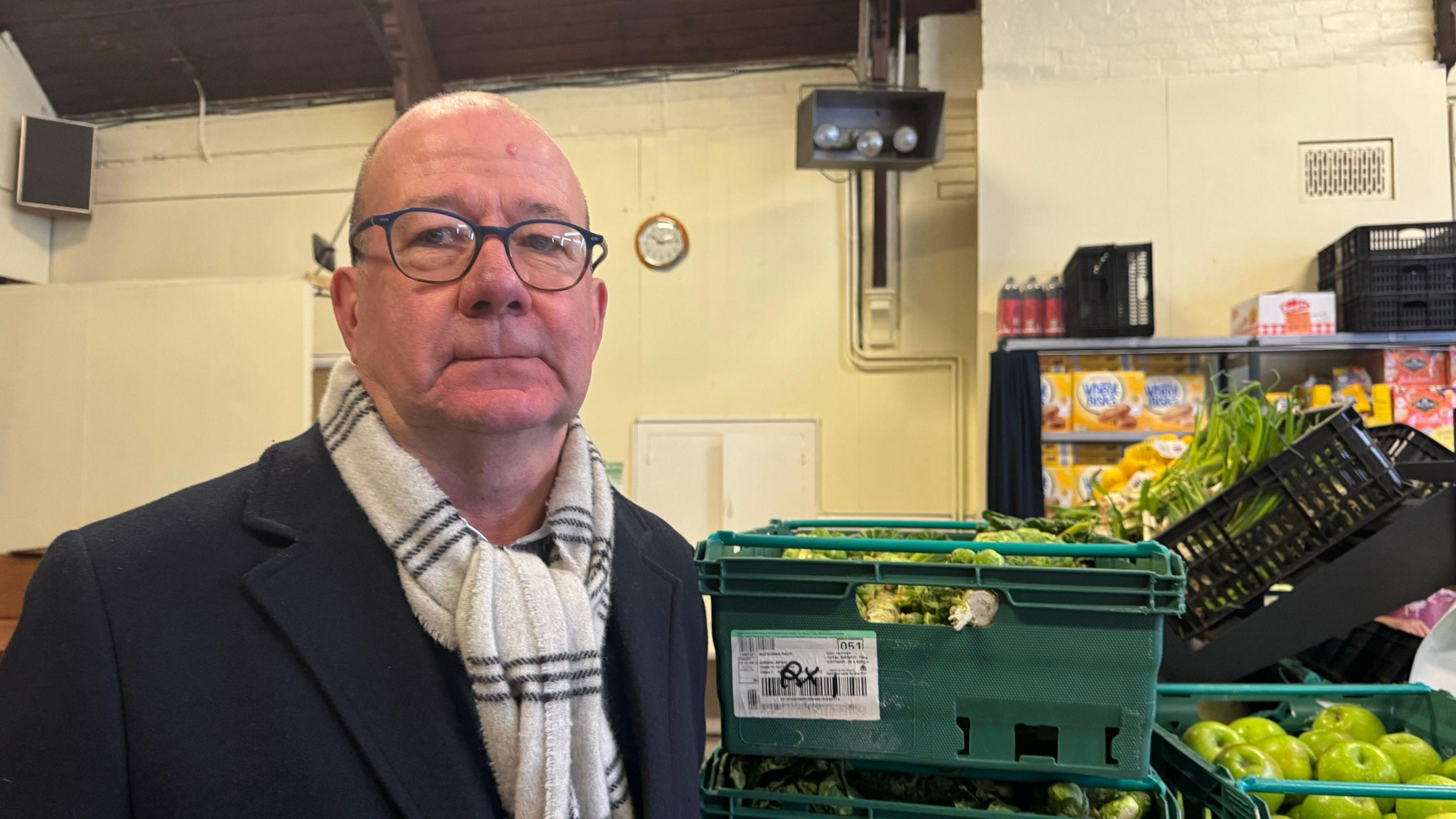 St George's Church warden Mark Ogden, a bald man wearing glasses, a black coat and a black and white scarf. He is stood next to green boxes filled with fruit and vegetables inside the church hall.