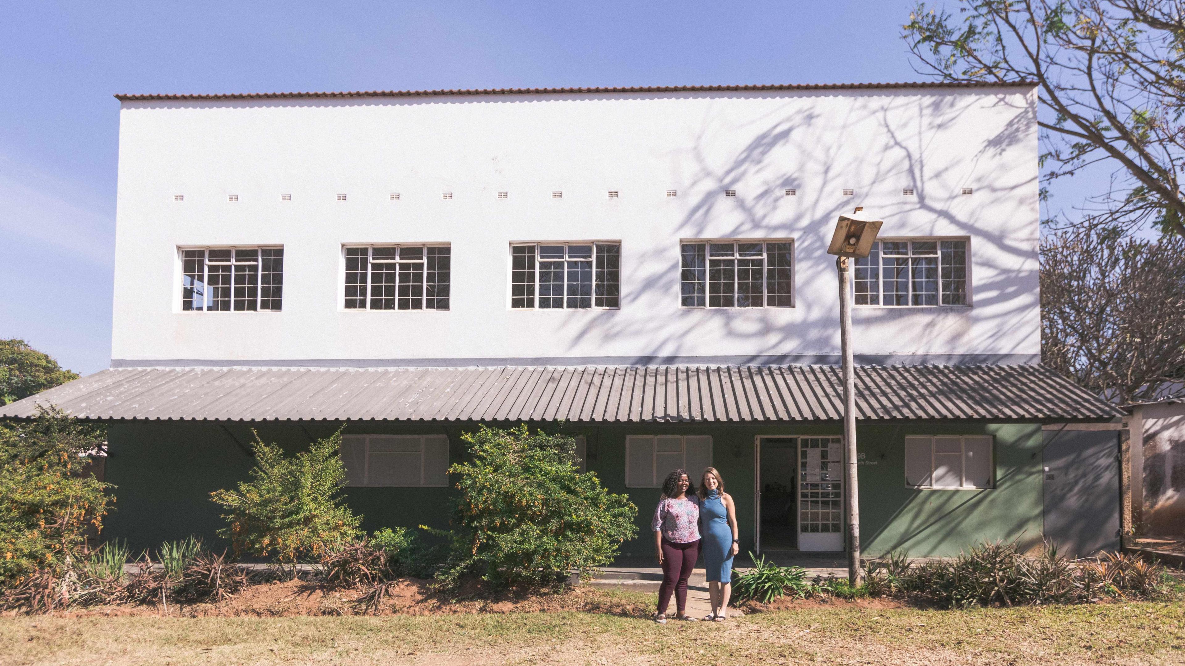 Two women stand outside a two-storey white building. There are five windows on the first storey, while a tin roof provides shade for a paved area outside the ground floor. 