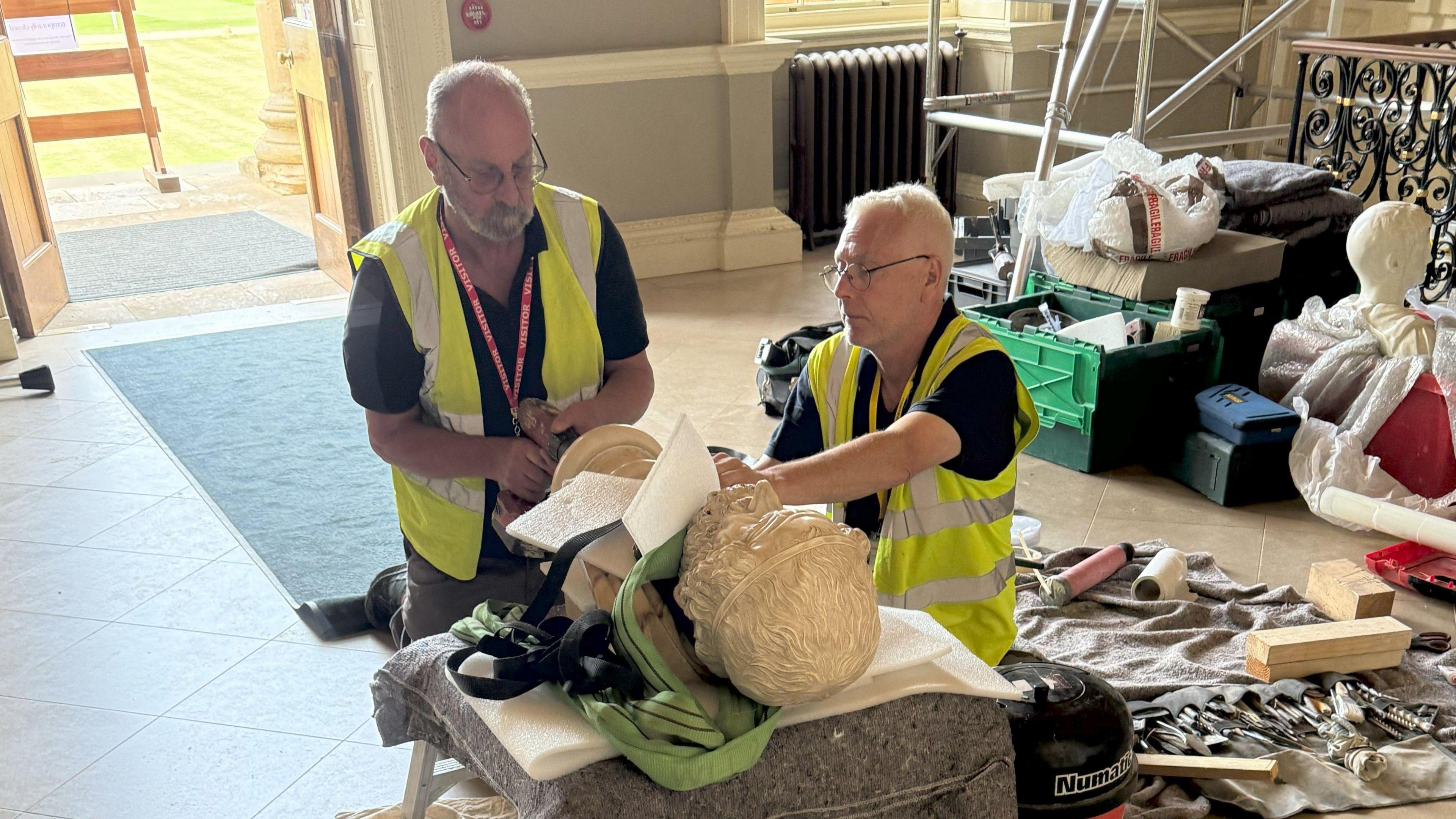 Two men in high-vis jackets working on a bust which is lying flat on a small table in a large room. They are surrounded by tools and scaffolding.
