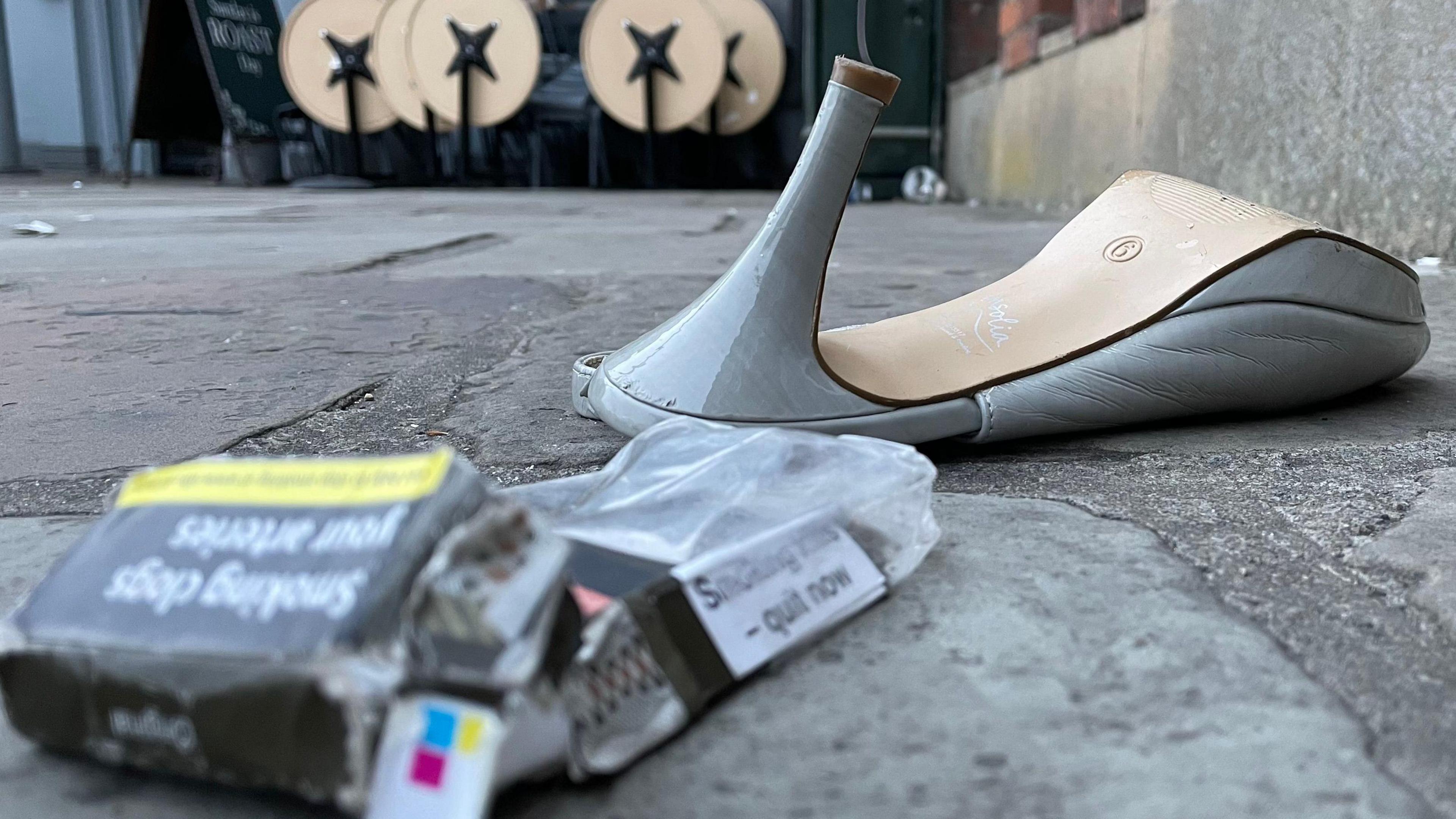 A discarded high-heeled shoe lies on a pavement next to an empty cigarette pack and small debris. In the background, outdoor tables are folded up outside a venue.