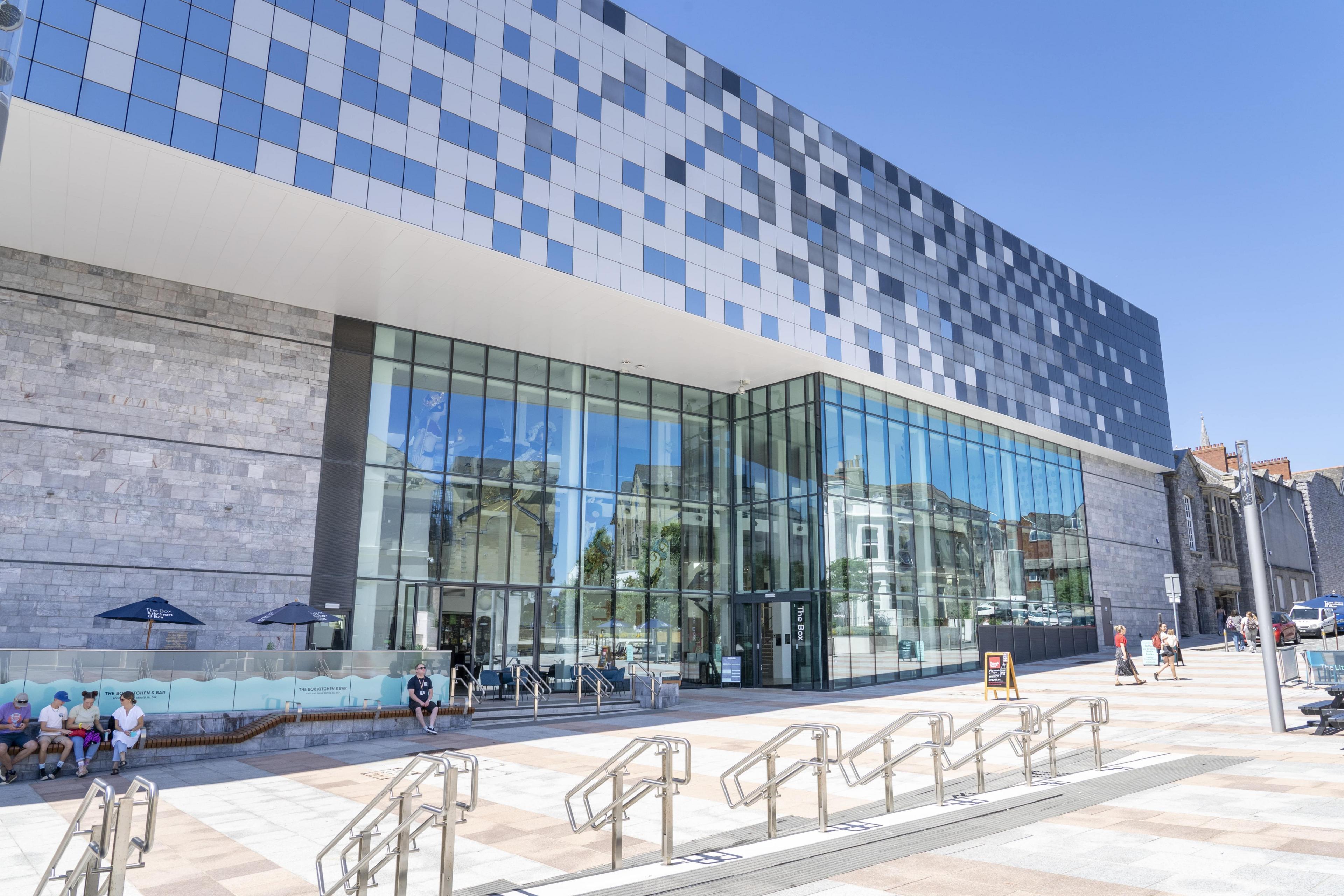 An exterior view of a modern public museum building, which has a large areas of glass and cladding that is like a grey and black chess board. There are people sitting outside the building. It is a sunny day, with bright blue sky. 