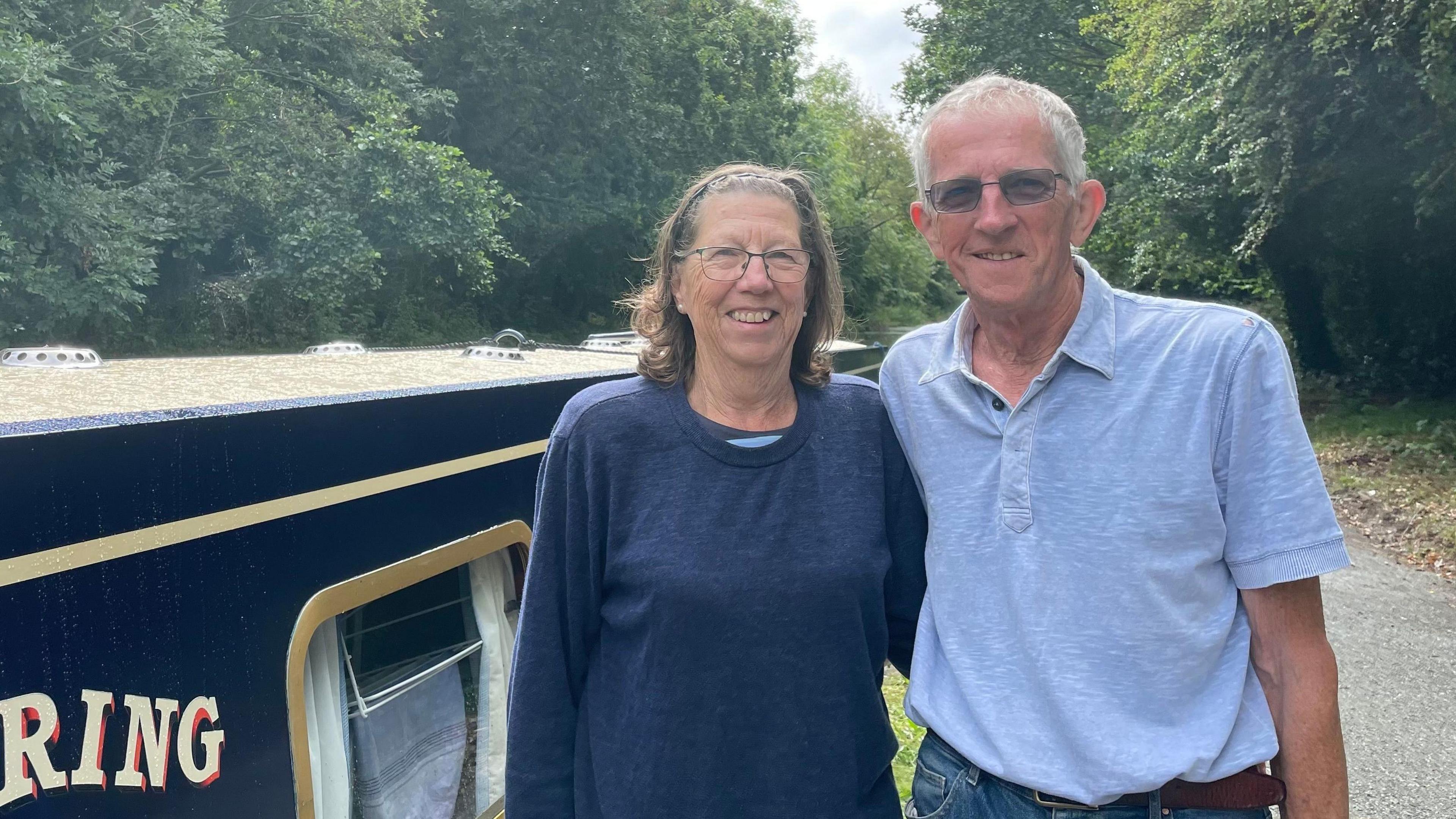 Bruce and Lauris Crook pose for a photograph beside a canalboat. They are both wearing glasses, she is in a blue crew-neck jumper and he is wearing a light-blue polo shirt