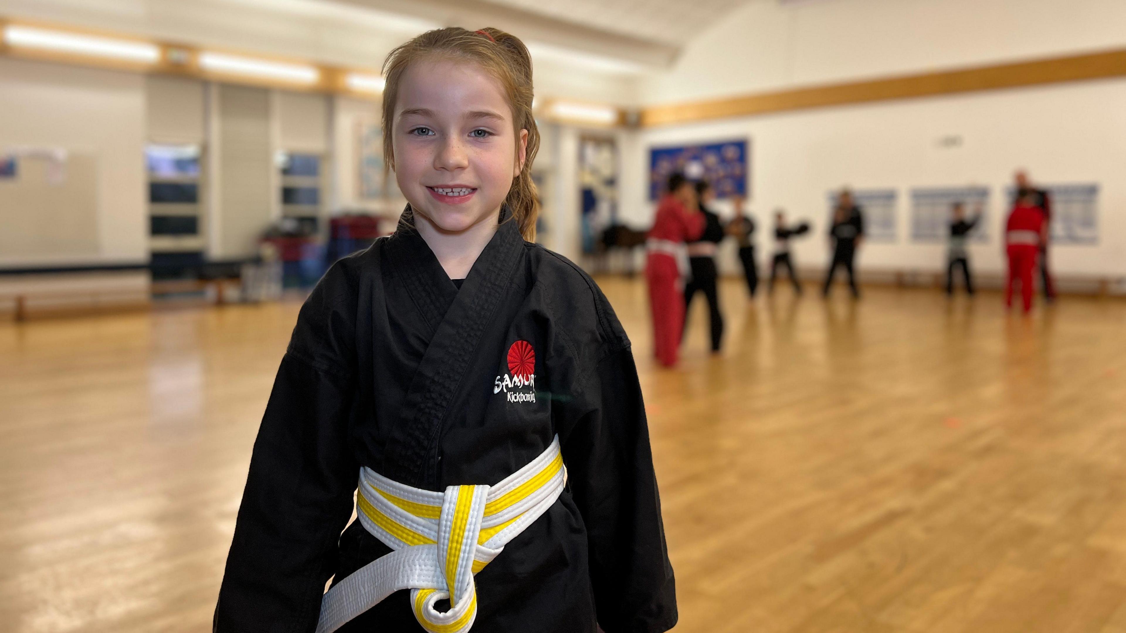 Evie smiles at the camera wearing a black gi with a Samurai Kickboxing logo on her chest. Children are practicing the sport behind her in the school hall. She has a yellow belt on which is loosely tied to her waist. She has light brown hair tied in a pony tail.