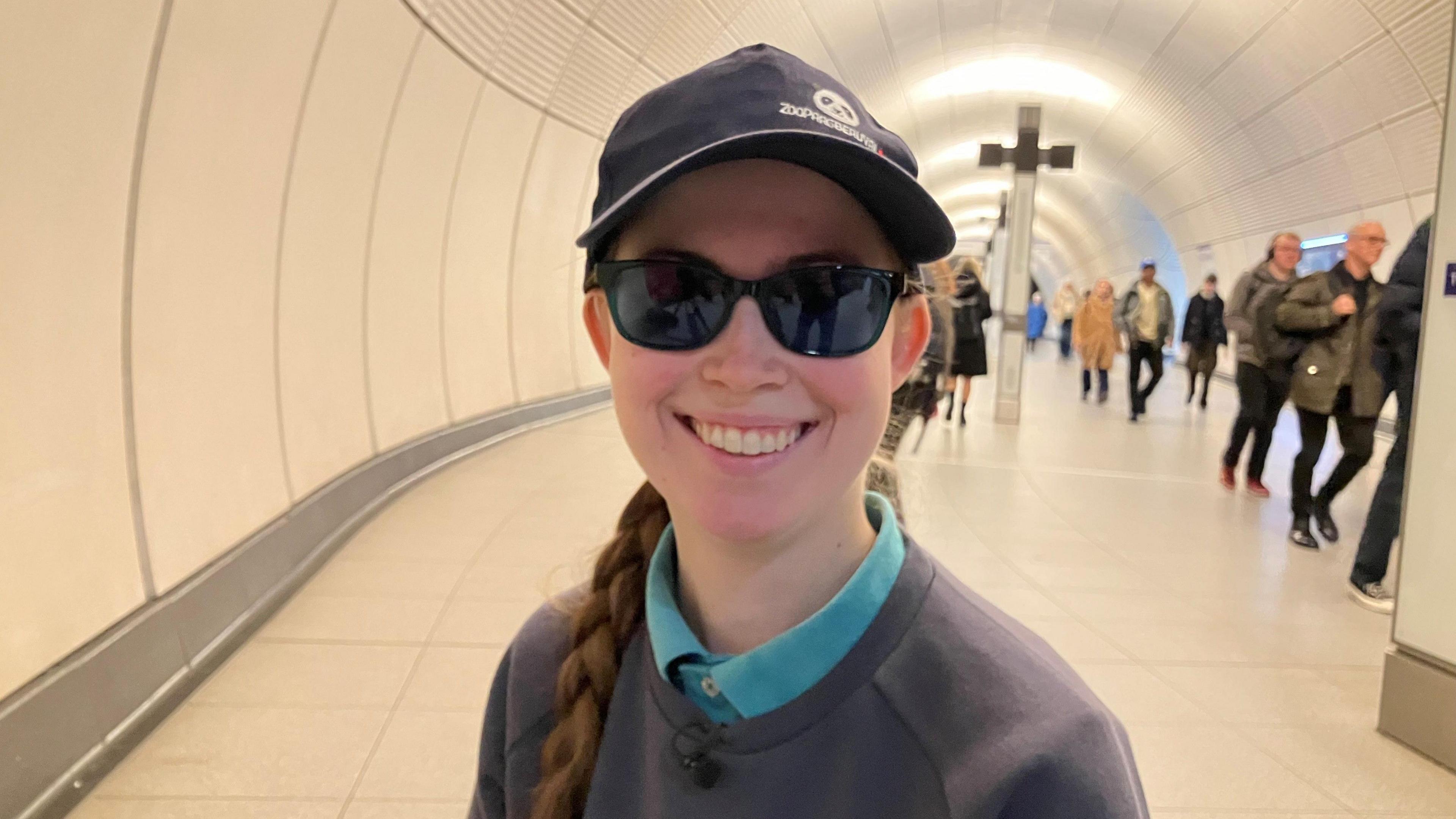 A woman with a long plait wearing a cap and sunglasses, a dark grey sweatshirt and a teal coloured polo shirt smiles at the camera. She is in a tiled tunnel on the London Underground.