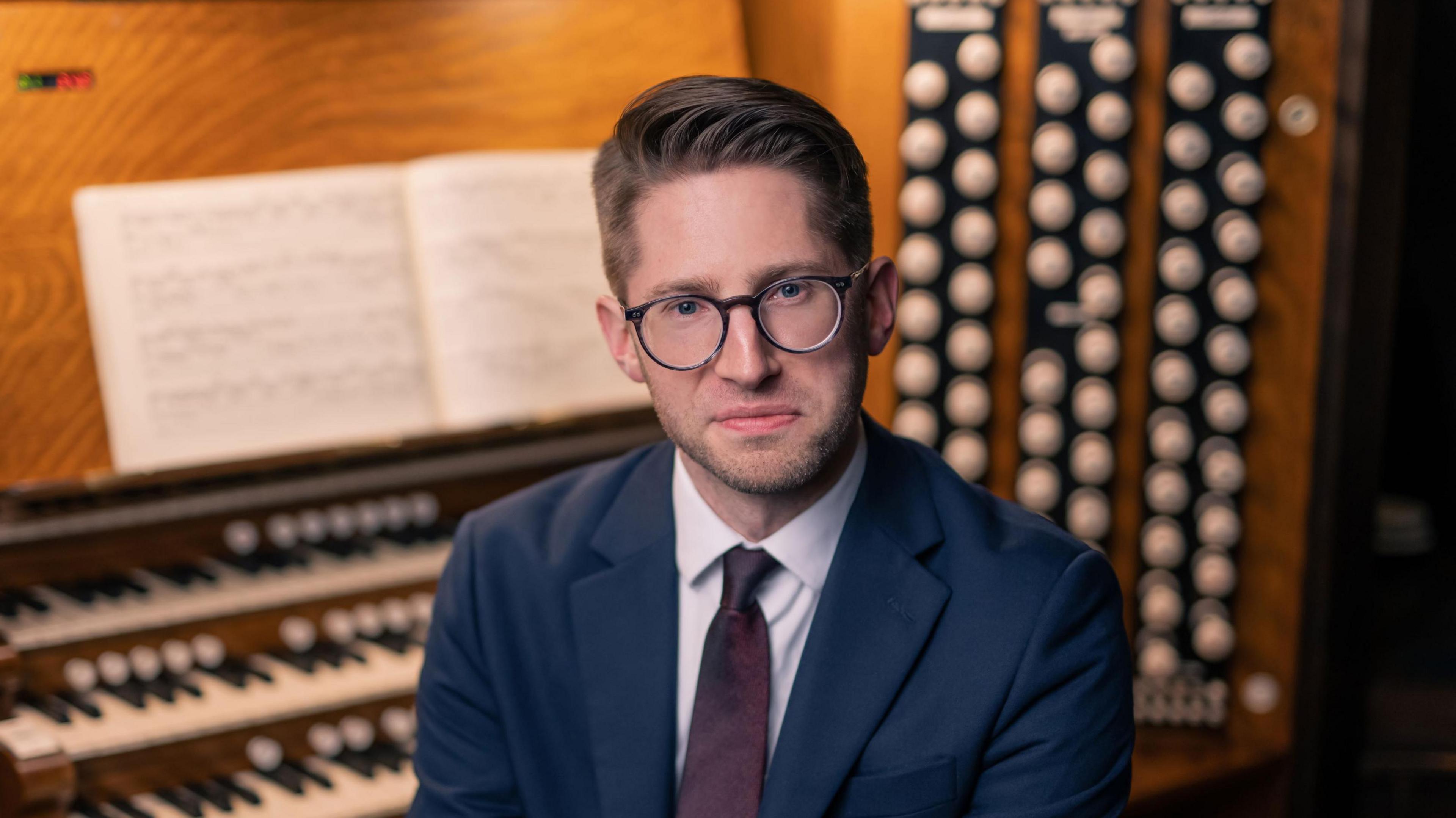 A man with shirt brown hair and glasses sits by a large wooden organ wearing a navy blue suit, white shirt and dark red tie.