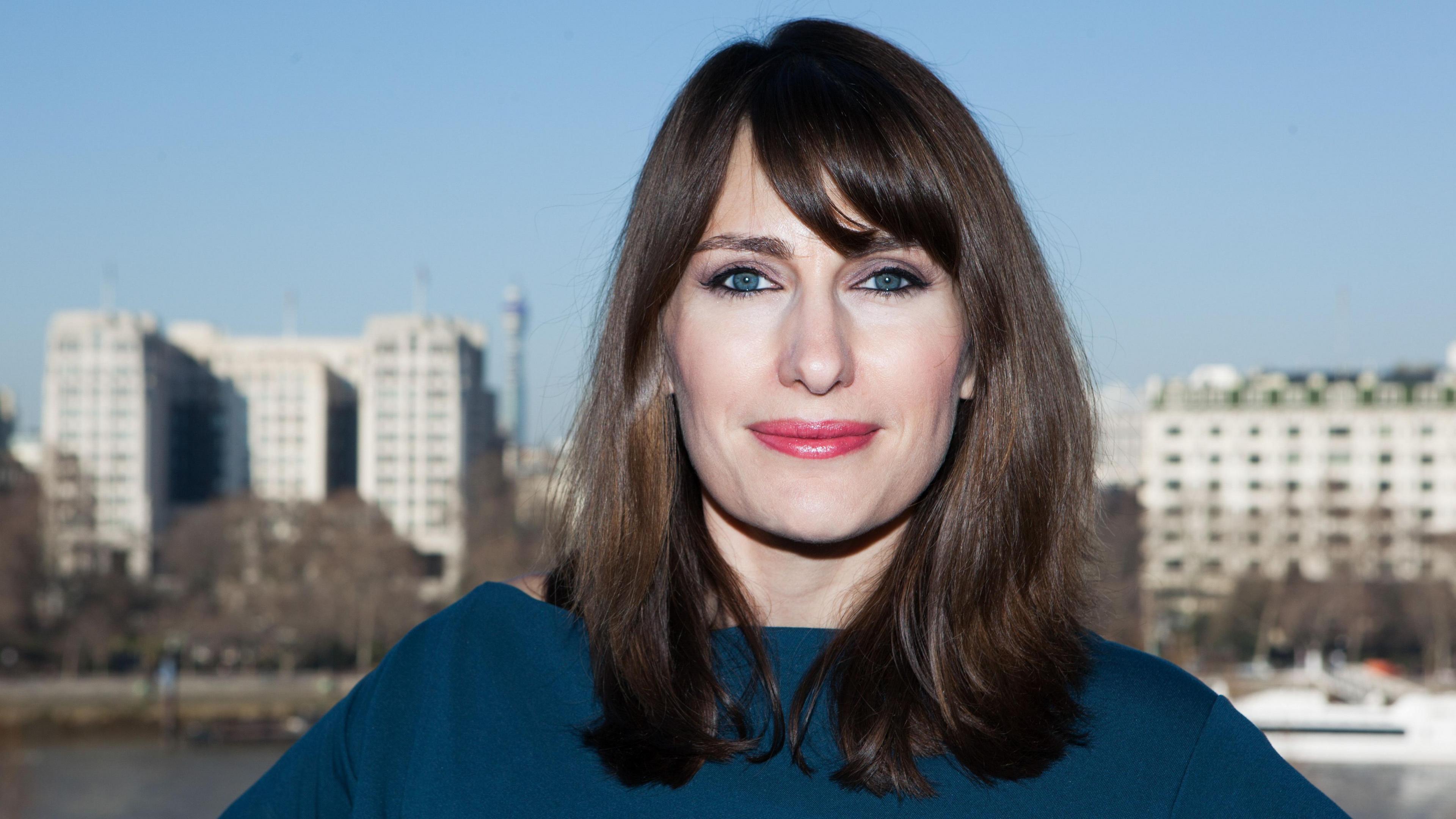 Alex O'Brien in a blue top. She has brown hair, a side fringe and wears pink lipstick. She stands in front of a river and high-rise buildings