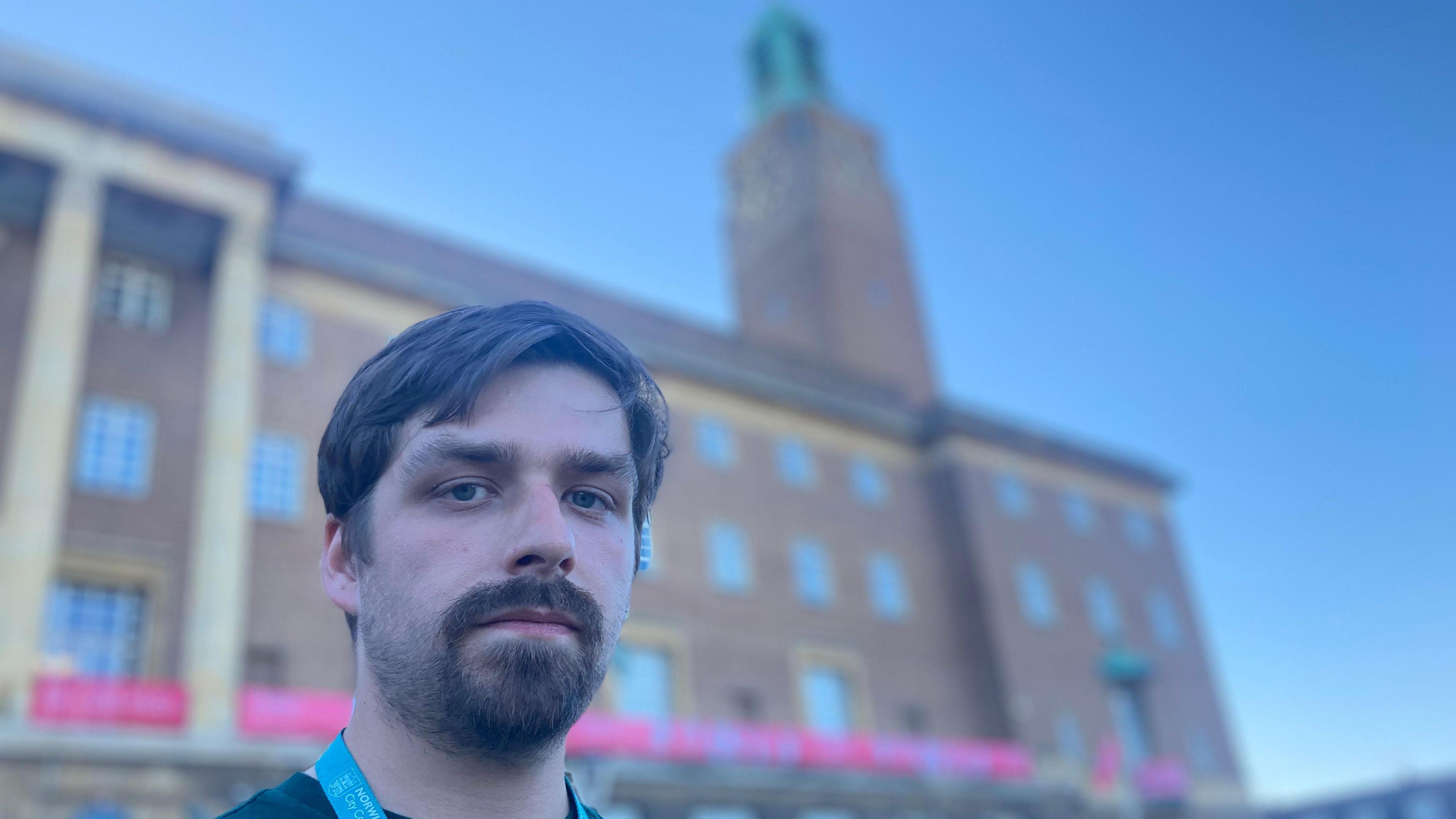 Alex Catt is looking directly at the camera. He is standing in front of the Norwich council building with a tall clock tower. He has a thick moustache and beard and a middle parting fringe.