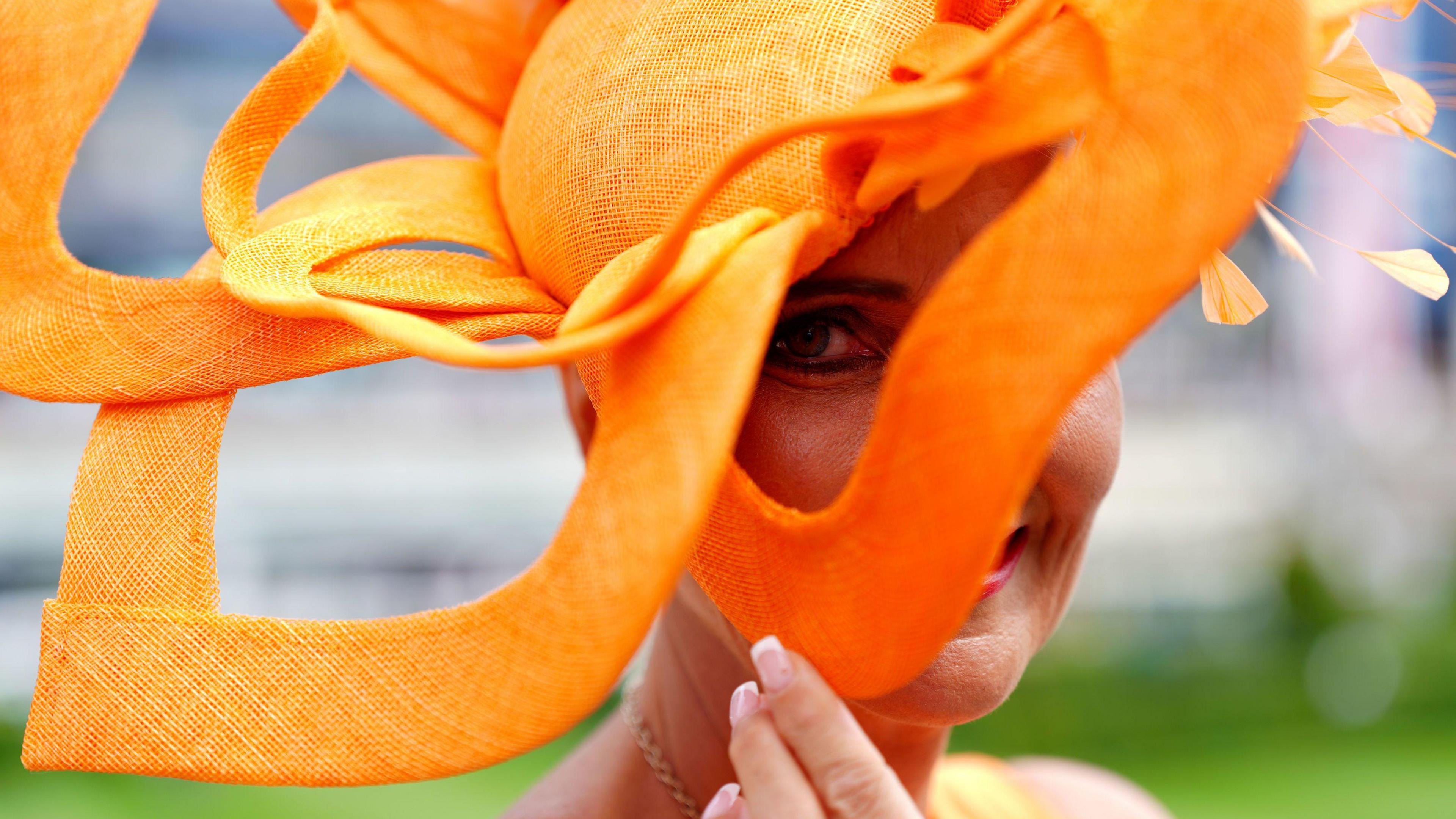 A woman wearing an orange fascinator looks through a gap in it at the camera. 