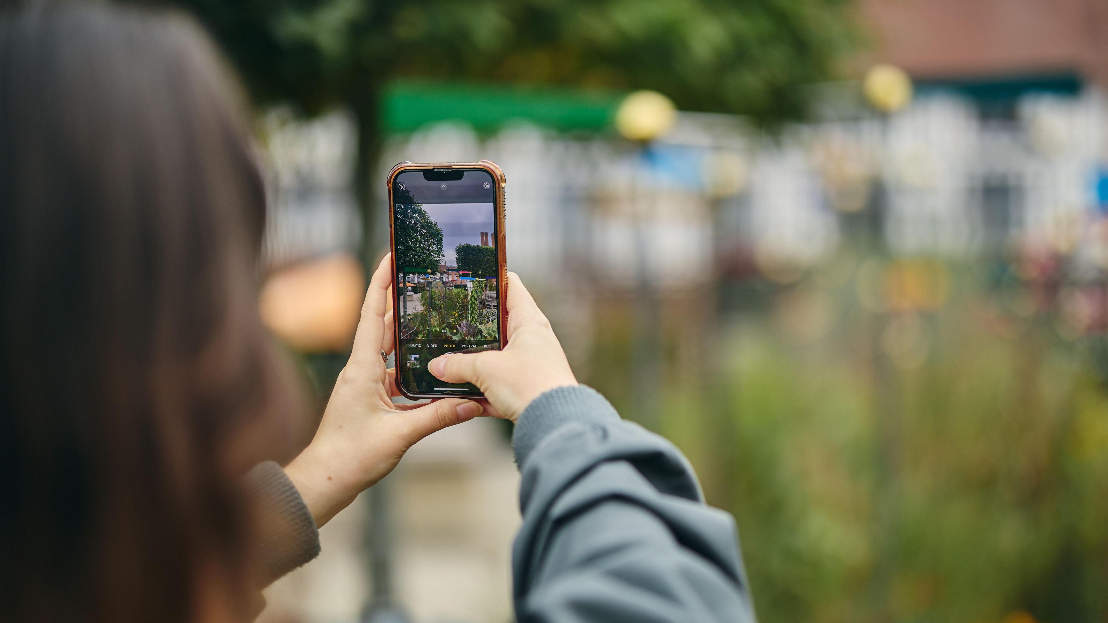 A woman holds a mobile phone up to take a picture of a scene in the background.