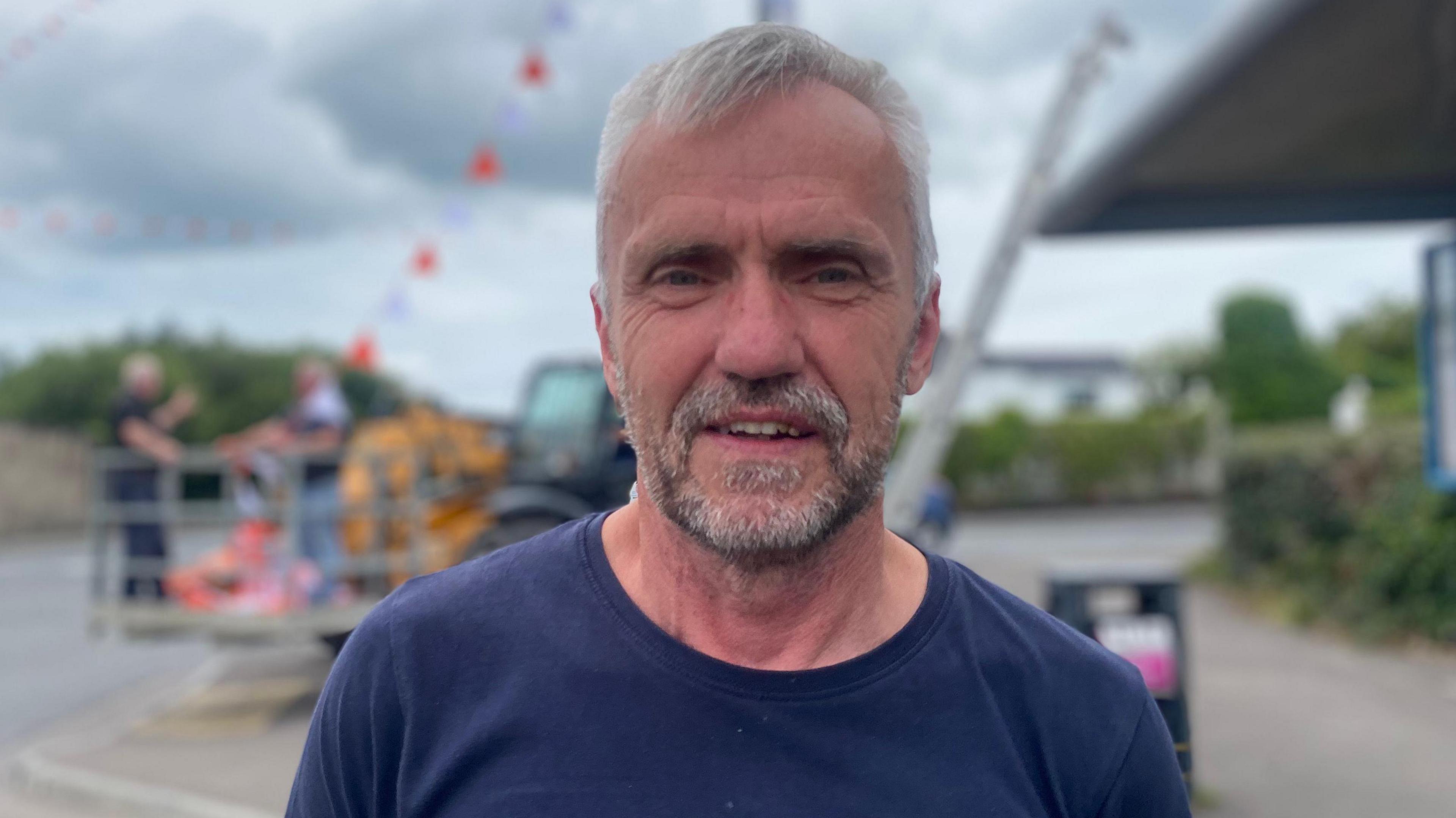 Peadar is looking directly into the camera wearing a navy blue t-shirt. The background of the photo is blurred but shows men erecting orange and white bunting.