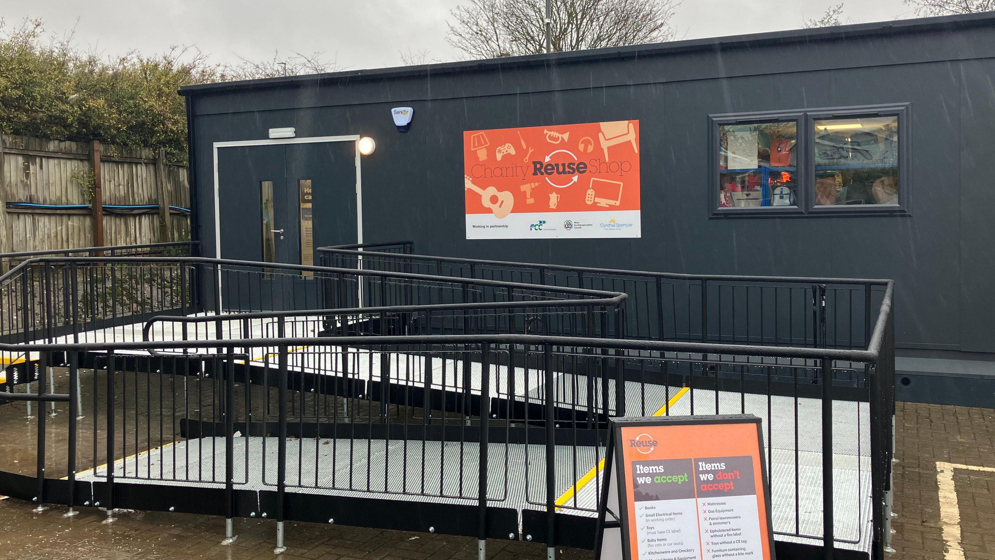 A anthracite flat-roof building with a silver aluminium ramp leading to the door. An orange reuse sign is on the side of the building.