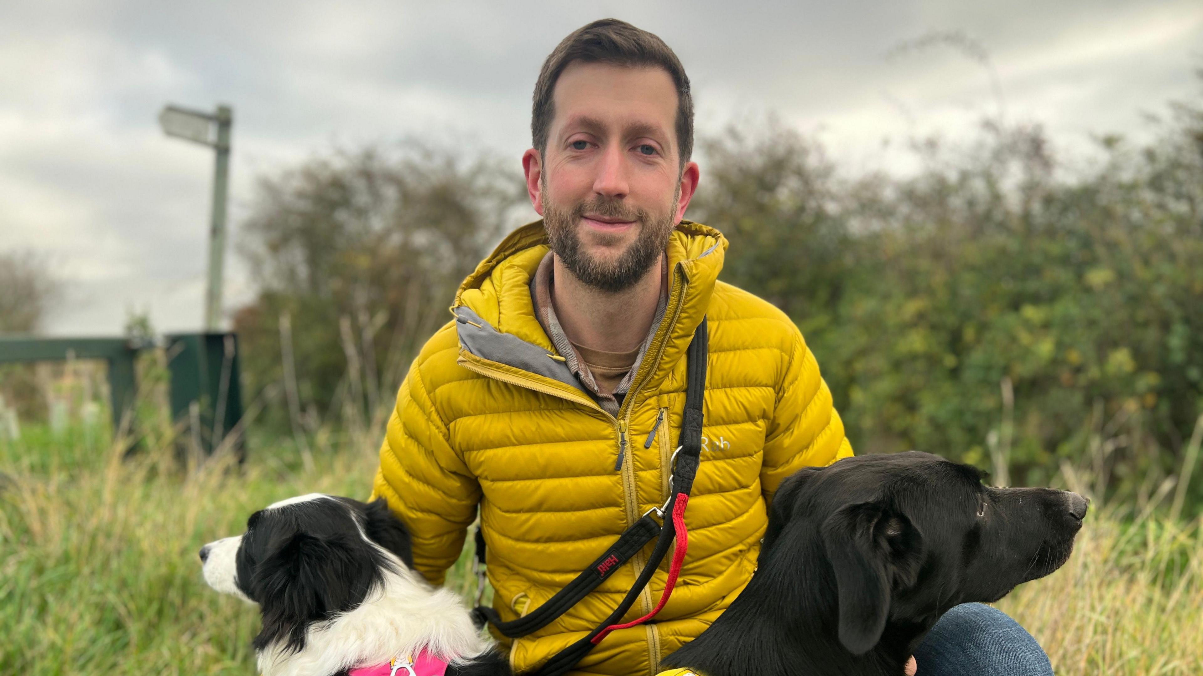 Mike Davey is holding his two dogs, and wearing a yellow puffer jacket. He is on a walk on a countryside footpath.