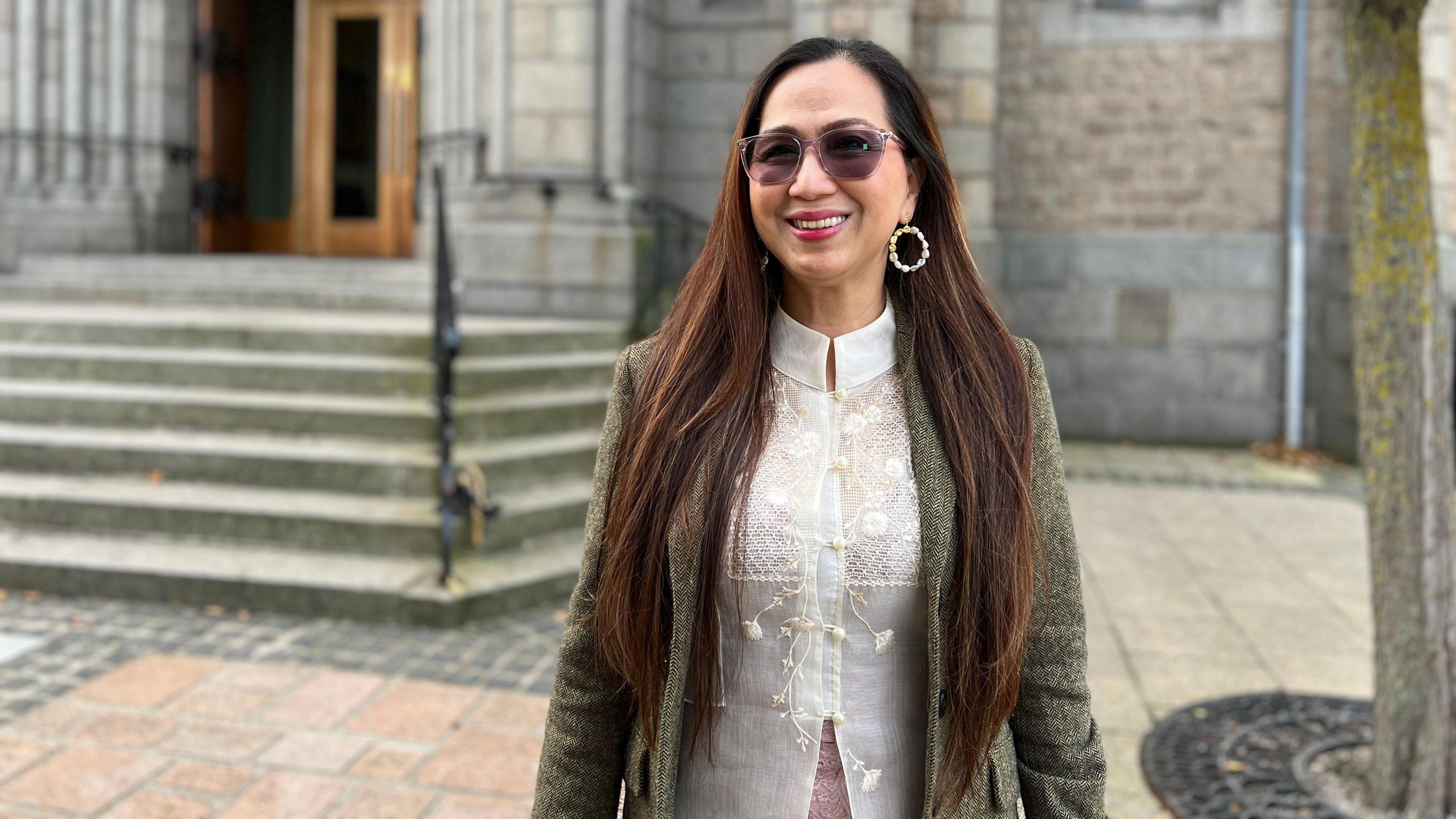 Marlene smiles at the camera outside the church. She is wearing a white blouse which has braided decorations on it and a light green jacket. She has hooped ear rings and sun glasses too. Her long dark hair comes down her chest.