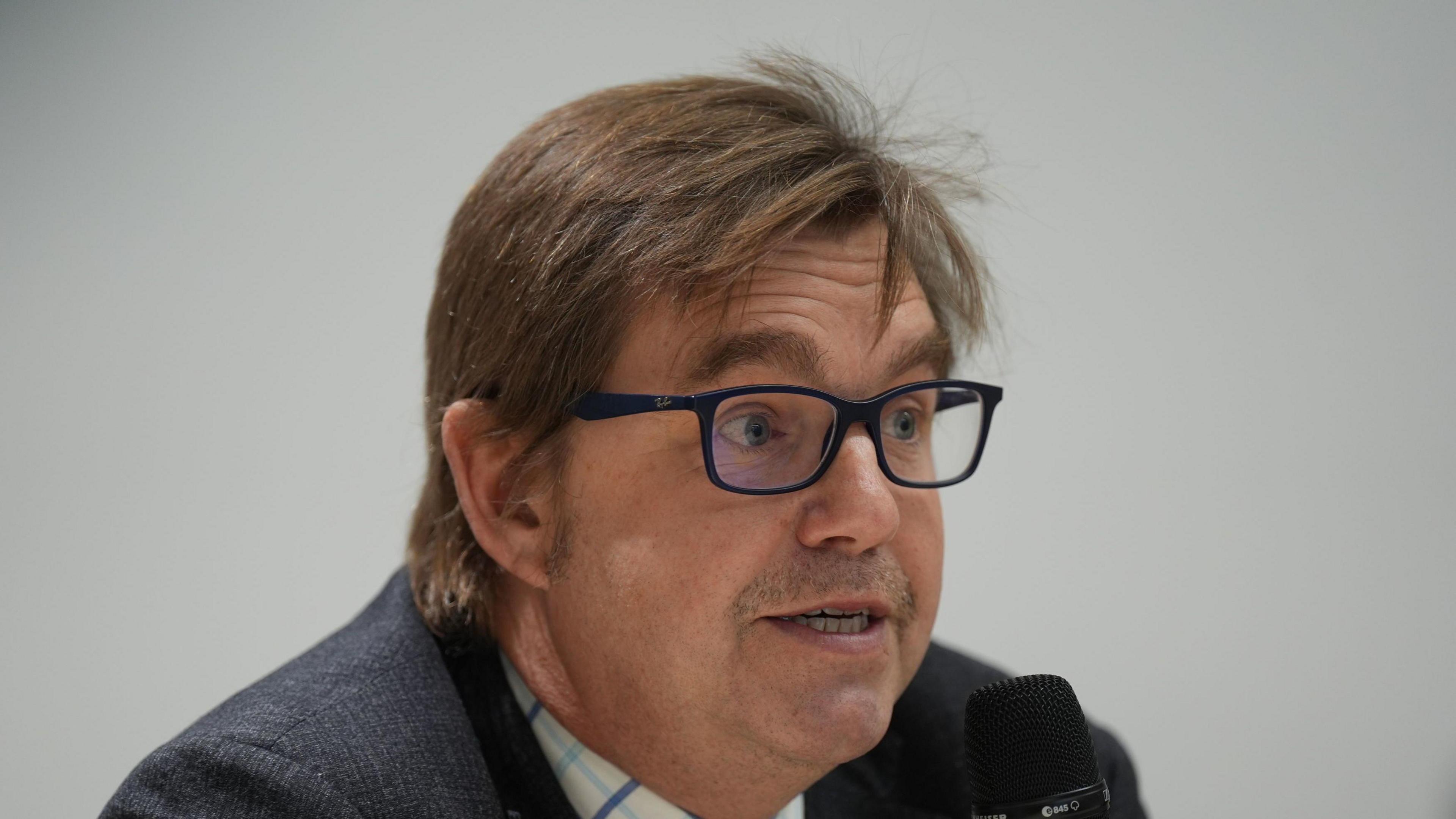 Nik Johnson is looking to the side of the camera while speaking into a microphone. He wears square-framed black glasses and a suit. He has a moustache and brown hair. 