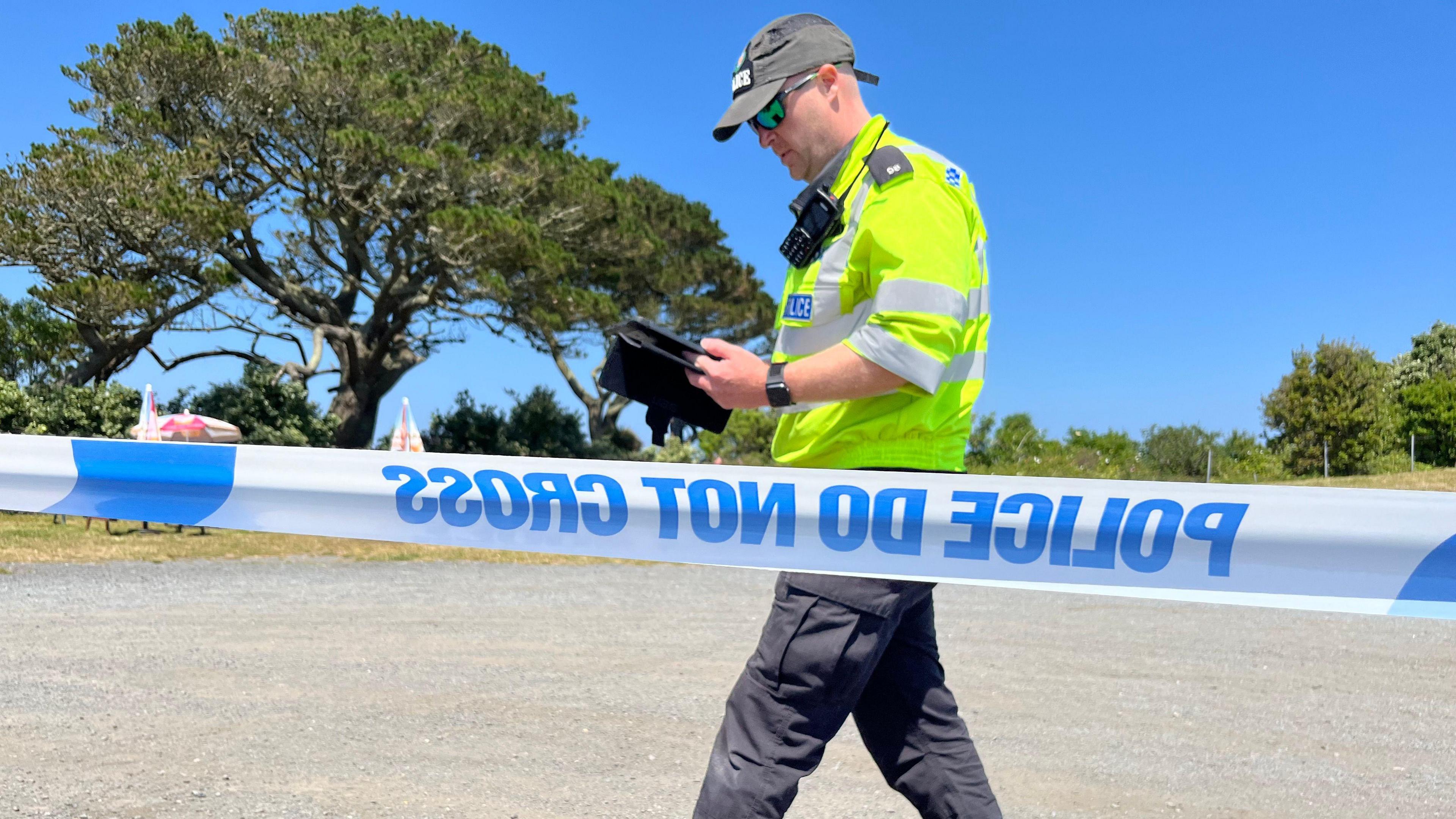 Image is taken behind blue and white police tape. In front of the tape a police officer wearing a high viz top and he is holding an electronic tablet.