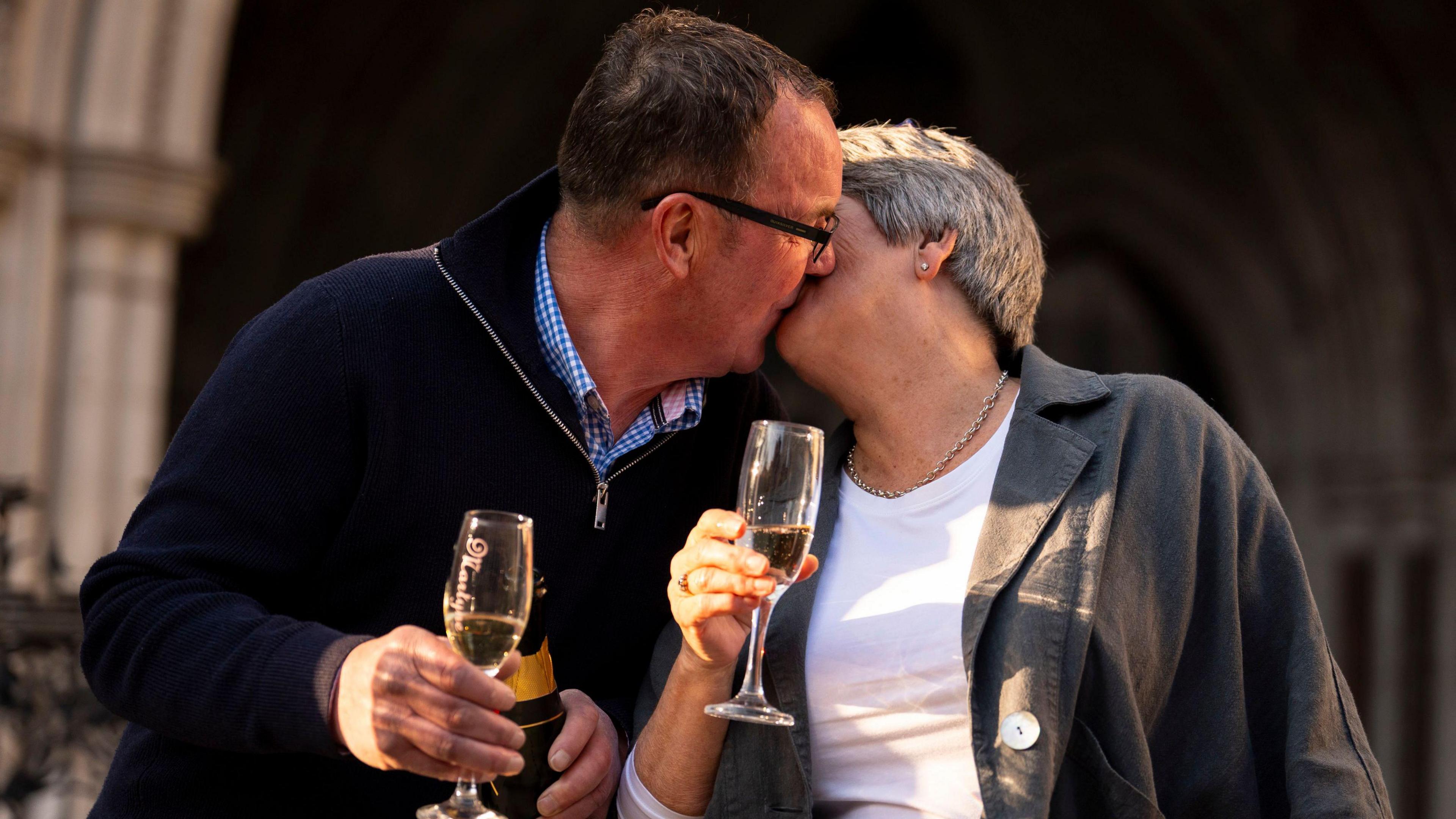 Corinne Durber with her husband Colin outside of the Royal Courts of Justice in London. They are kissing each other on the lips as they hold champagne flutes in their hands.