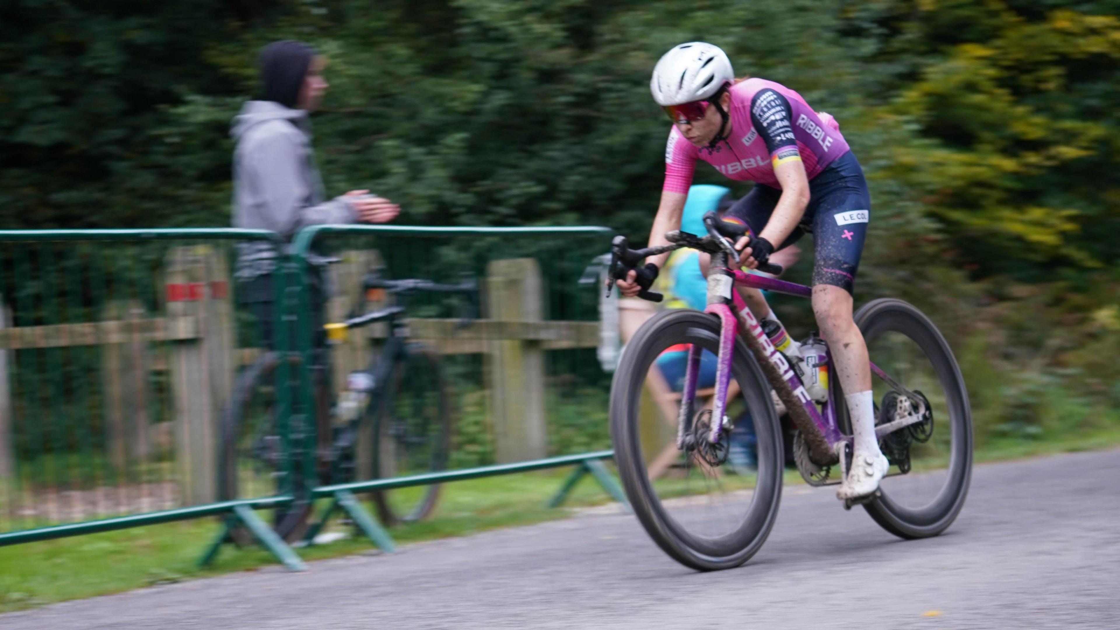 Sophie Wright riding a gravel bike at speed during a race; she has mud on her legs and bike. A spectator in the background stands behind railings clapping.