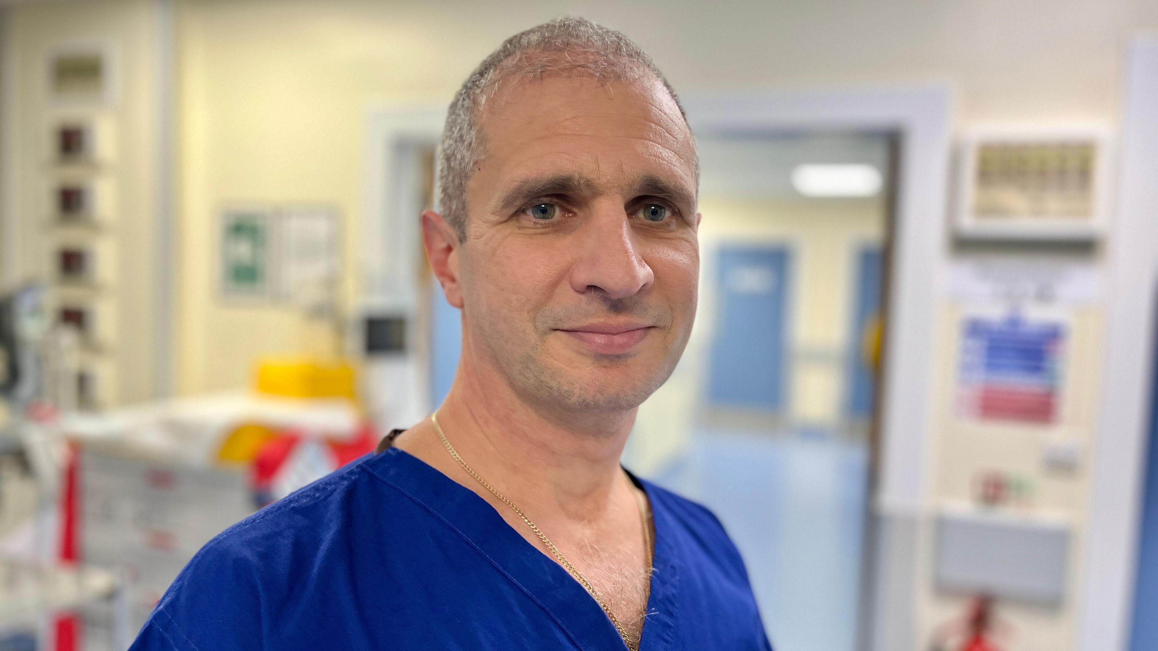 A man wearing blue surgical clothes staring at the camera. He has short grey hair and is standing outside an operating theatre